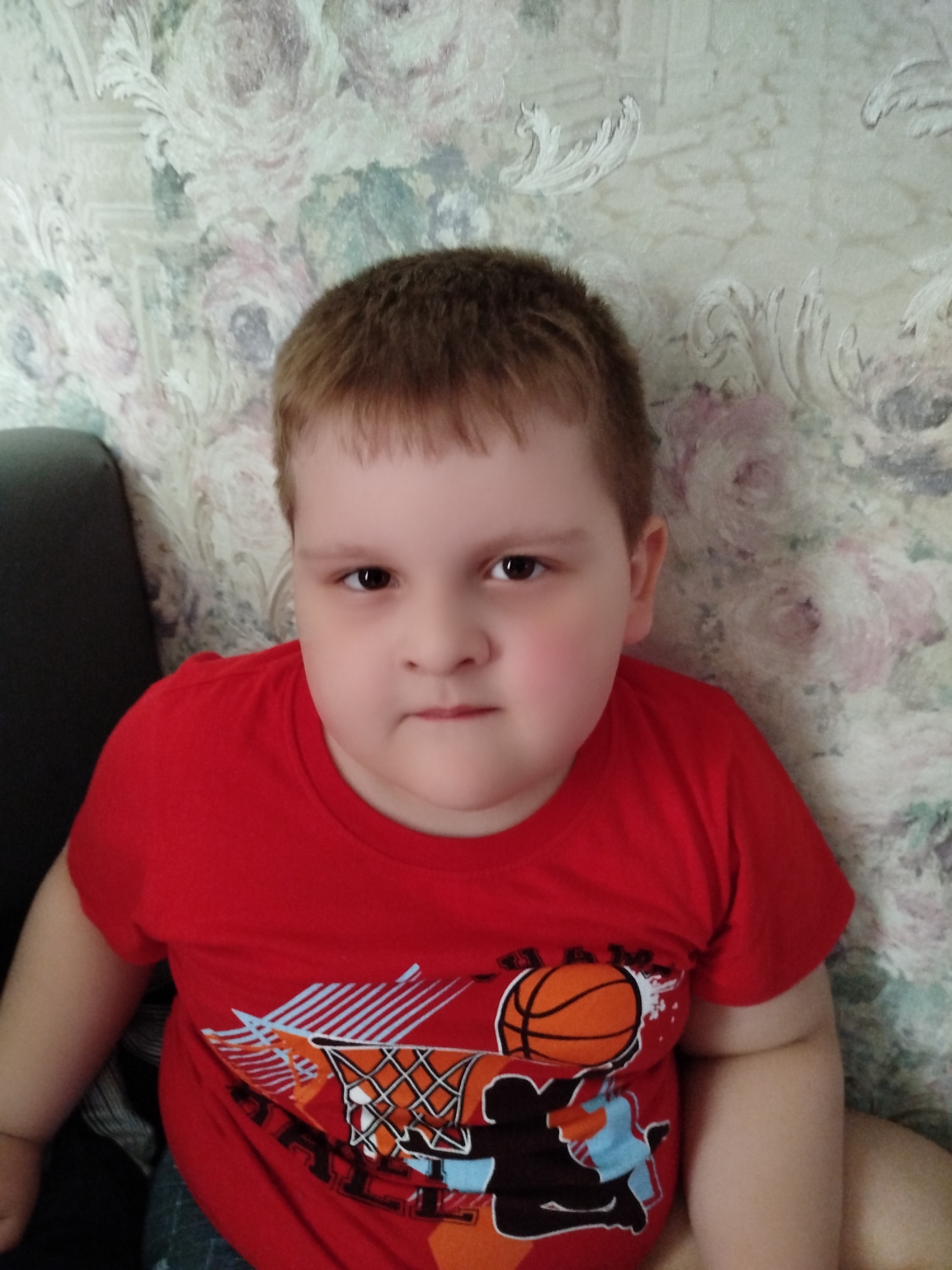 A young boy with short brown hair, wearing a red t-shirt with a basketball graphic, is pictured sitting in front of a floral patterned wallpaper. The boy's gaze is directed towards the camera. The photograph appears to be taken indoors, likely in a residential setting. No specific location in Saratov, Russia, can be identified from the image itself.