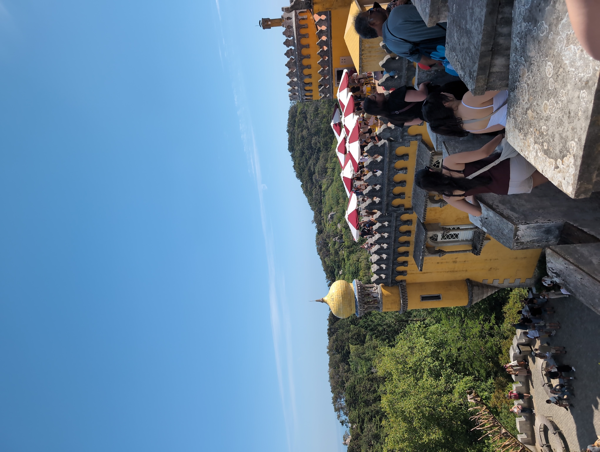 The image shows an outdoor scene in Sintra, Portugal. The prominent feature is a section of the Pena Palace, with its yellow walls and decorative architectural details. The sky is blue. People are visible on the palace's terrace and in the surrounding areas, suggesting they are tourists or visitors. The landscape around the palace is characterized by lush green vegetation, including trees. Red and white awnings are visible near the crowd.