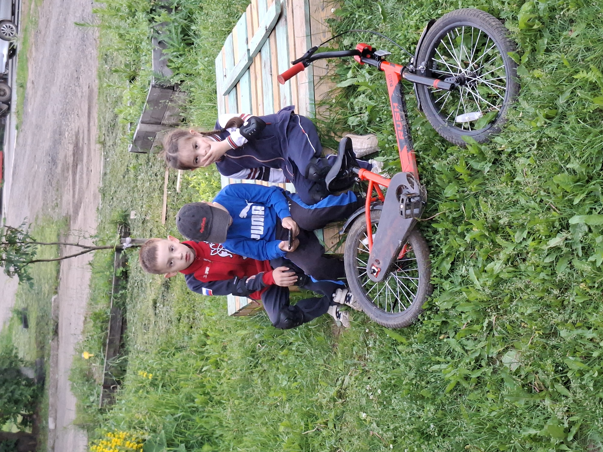 In Votkinsk, Russia, three children are seated on a wooden platform outdoors. A young boy on the left wears a red and black tracksuit and black pants. In the center, another boy in a blue Puma sweatshirt and black cap looks down at his phone. To the right, a young girl in a dark blue tracksuit with white stripes covers her mouth with her hand. A red BMX bicycle lies on the grass in the foreground, with its front wheel positioned to the right. The children are surrounded by green grass and some yellow wildflowers. A wooden structure is visible behind them, and a dirt road with some distant vehicles is in the background.