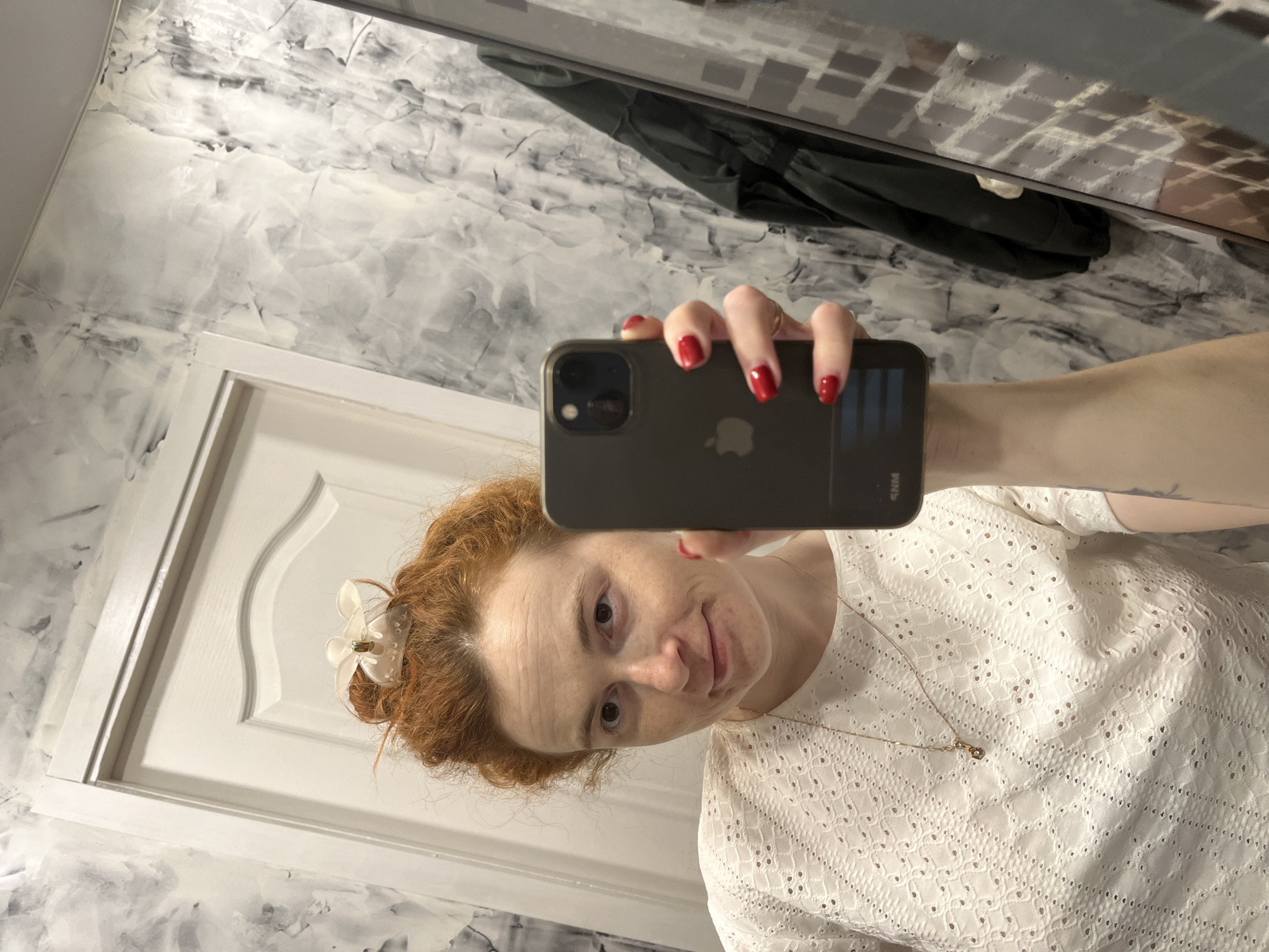 A woman with red hair is taking a selfie in a bathroom with grey and white marble-patterned walls and a white door. She is wearing a white lace top and a necklace, and has a white flower clip in her hair. Her nails are painted red. The phone she is holding is black and has an Apple logo. A black jacket is visible in the mirror's reflection. The location is likely a private residence rather than a public space in Chaikovsky, Russia.