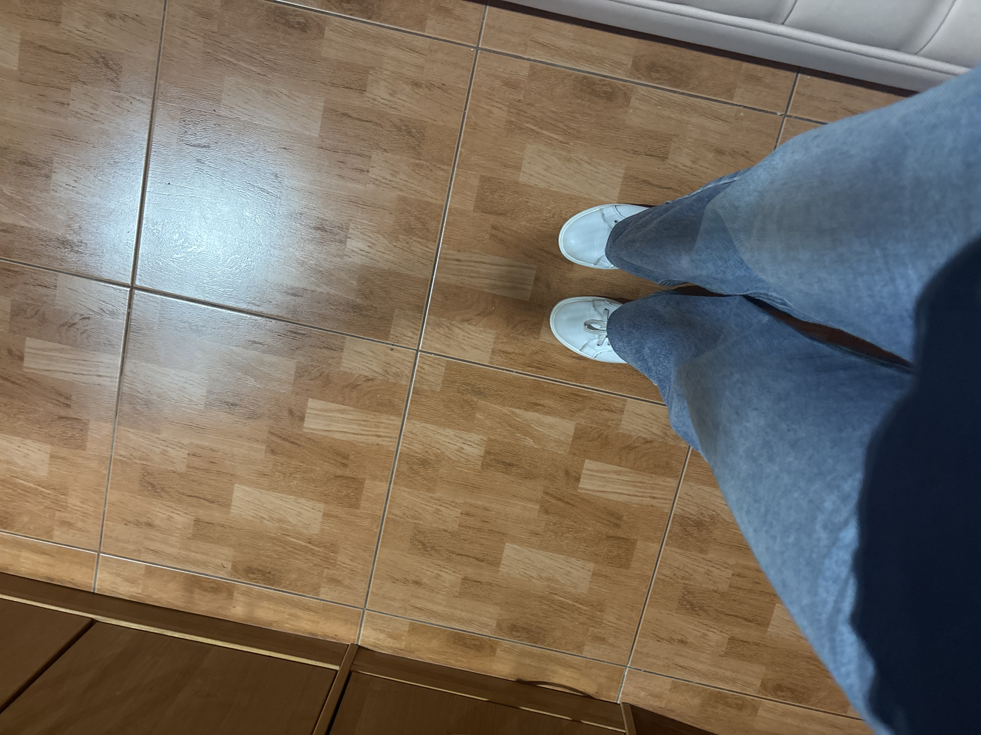 A high-angle shot reveals a section of tiled flooring, light brown with a wood-grain pattern. A pair of legs clad in light blue jeans, extending from the knees down, are positioned on the right side of the frame. The feet are shod in white sneakers. The top edge of a white object is visible at the top edge of the frame. A portion of what seems to be wooden furniture is at the bottom-left corner of the image. The location is Ферма, Russia.