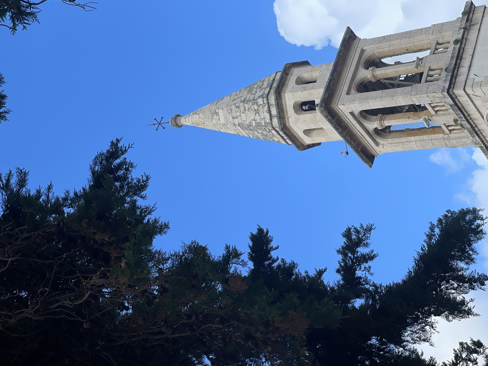 The photograph presents a low-angle view of a stone church tower rising against a bright blue sky interspersed with a few clouds. The tower is constructed of light-colored stone and features a pointed spire topped with a metal ornament. The tower's architecture includes arched openings, some of which contain bells. Dark green foliage from trees frames the lower part of the image, adding a contrasting element to the scene. The location is Unknown, Unknown.