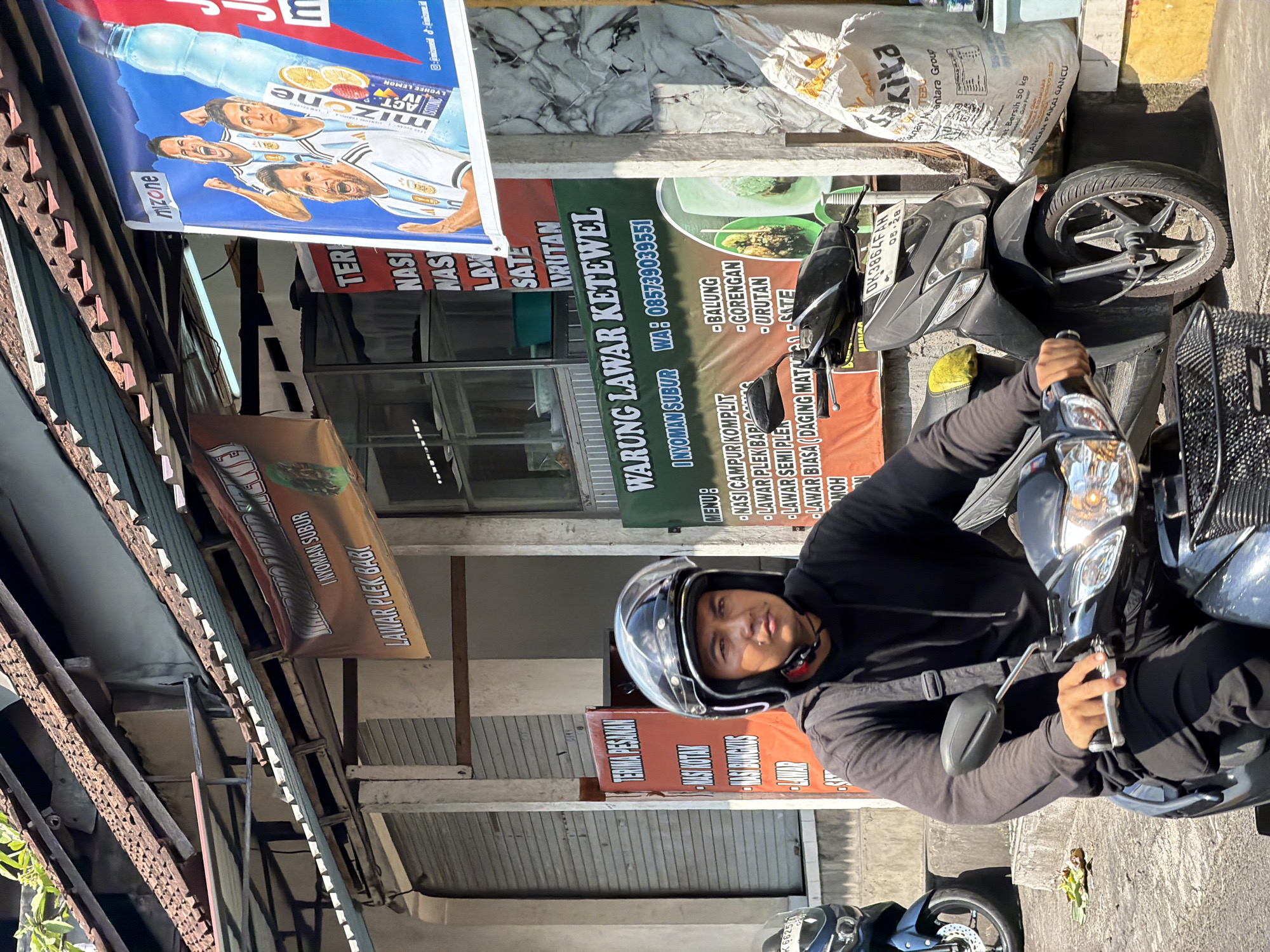 This media file captures a street scene in Ubud District, Indonesia, likely during daylight hours as indicated by the strong shadows. The primary subject is a person wearing a black hoodie and a motorcycle helmet, seated on a dark-colored motorcycle. The person is looking towards the camera with a neutral expression.
The setting appears to be a roadside stall or shop, with various banners and signs prominently displayed. One large banner advertises "mizone" with images of athletes and the flavors "LYCHEE LEMON" and "ACT IN". Another banner, predominantly green, reads "WARUNG LAWAR KETEWEL" and lists menu items like "BALUNG", "GORENGAN", "URUTAN", and "SATE". It also provides contact information: "I NYOMAN SUBUR WA: 085739039551".
Other visible signs include "TERIMA PESAMAN", "NASI BUNGKUS", and "LAWAR PLEK BAG". A large sack labeled "Sakita" and "antara Group" is also visible, suggesting a commercial area. The motorcycle itself has a license plate that reads "DK3864FAH 05-28". The overall scene is a bustling street environment with an emphasis on food vendors.