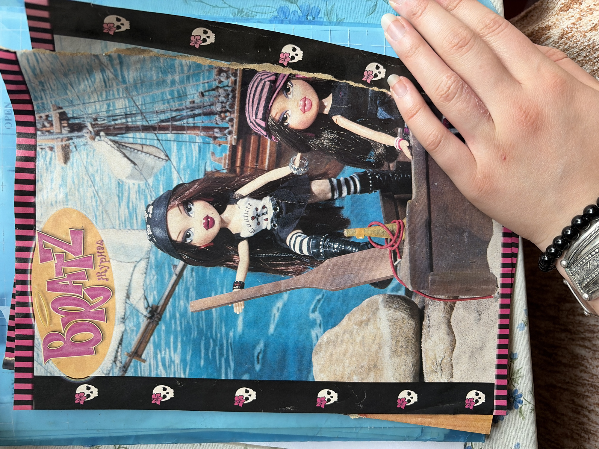 The image shows a close-up of a magazine or book open to a page featuring Bratz dolls. The dolls appear to be pirate-themed. One doll wears a black bandana, a black top, a black skirt, and striped thigh-high socks with black boots. She holds a wooden oar. The second doll, partially obscured by the first, wears a pink and black striped bandana, and a black top. Both dolls have large eyes and full lips, characteristic of the Bratz doll brand. The background of the page depicts a seascape with a sailing ship. Decorative elements on the edge of the page include black strips with repeating patterns of skulls with pink flowers and pink and black stripes. A human hand with light skin and short fingernails is visible in the lower right portion of the image, touching the edge of the page. A beaded bracelet is worn on the wrist. The image appears to be taken indoors, with a textured, possibly wooden surface visible at the bottom and a blue-patterned fabric beneath the magazine. The location of Malyshevo, Russia, is not depicted or suggested in the image.