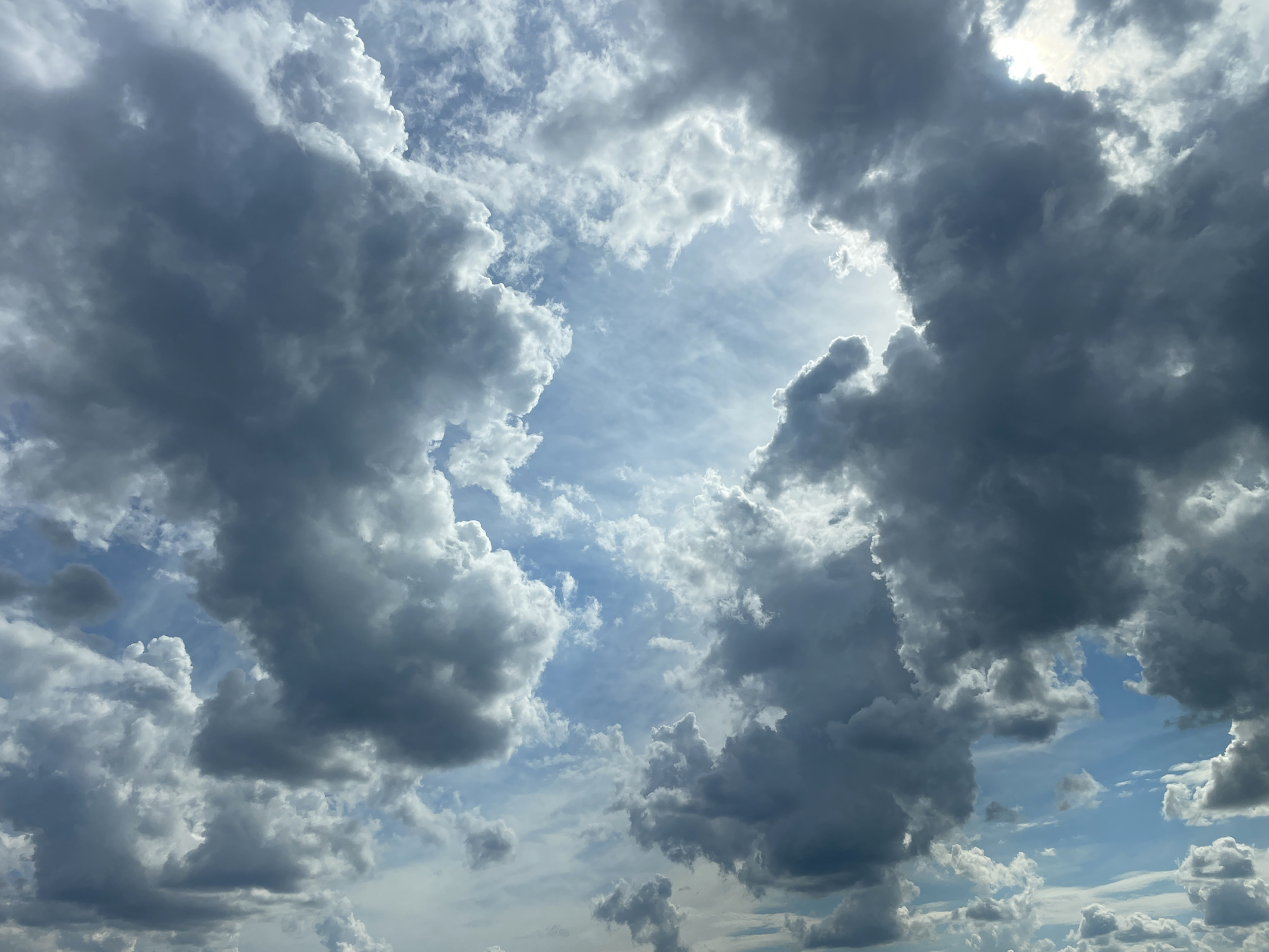 A low-angle shot shows a dramatic sky filled with a mix of dark, heavy storm clouds and lighter, more wispy clouds. The sun is partially obscured by the clouds, creating bright highlights and casting a diffuse light. The blue sky peeks through in several areas, particularly in the upper center of the frame. The overall impression is one of an overcast or approaching storm, with a sense of dynamic movement in the cloud formations. The image is an outdoor scene, likely taken during daylight hours. There are no discernible landmarks, people, or specific objects that would allow for the identification of a specific location within Comrat, Moldova, or any other city.