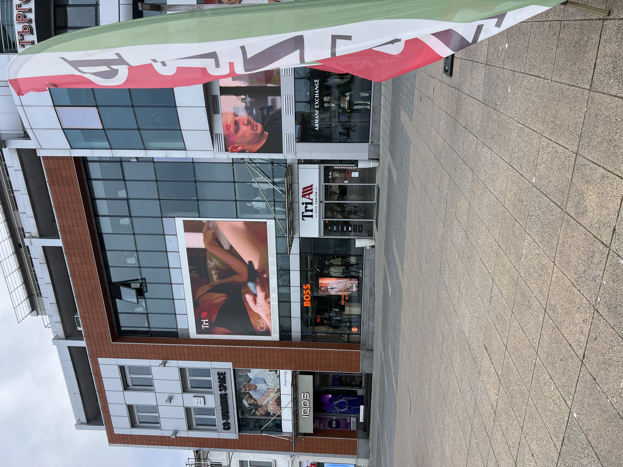 The image shows the facade of a multi-story commercial building, likely located in a shopping district in Burgas, Bulgaria. The building features a mix of modern architectural styles, with large glass windows and brown tiled sections. On the ground floor, several storefronts are visible, including Armani Exchange, TriAll City Centre, BOSS, and an IQOS store. Digital billboards display advertisements for brands like Calvin Klein and Hilfiger. A banner is partially visible at the top of the frame, obscuring the top portion of the building. The street in front of the building is paved with stone tiles. A Co-Working space is also advertised.