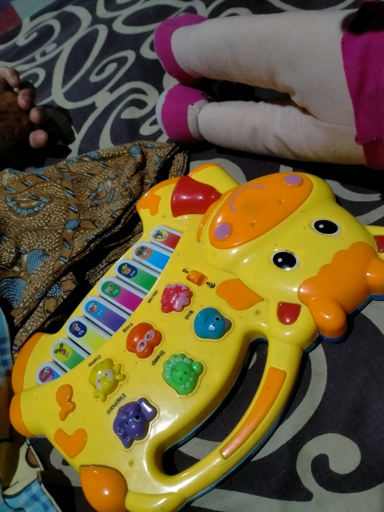 A yellow toy giraffe with a musical keyboard and animal buttons is placed on a dark patterned blanket. The toy has a smiling face with orange accents, black eyes, and pink circles on its head. The keyboard features colorful keys with pictures of animals and text labels such as "Frog," "Cattle," and "Sheep." To the left of the toy, a person's hand, with visible fingers and knuckles, rests on a piece of patterned brown fabric. In the background, a portion of a large, light-colored stuffed toy with pink accents is visible, suggesting it is lying down. The setting appears to be indoors, likely in a home environment. The location of Playen, Indonesia, is not discernible from the image itself and is provided as external context.