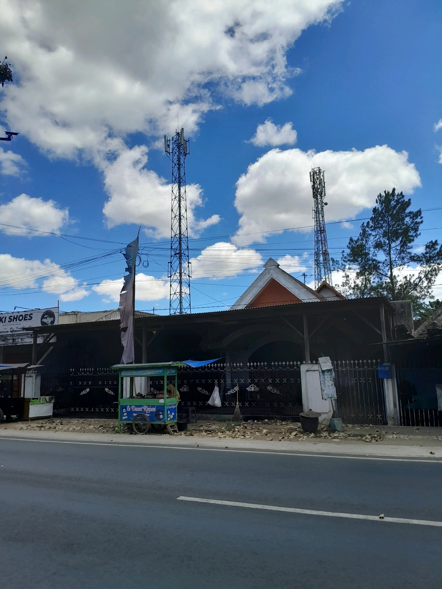 The image shows a street scene in Talangagung, Indonesia, under a bright blue sky with scattered white clouds. In the foreground, a paved road is visible, with a white dashed line marking the lane. Along the side of the road, a small, green cart with a striped awning is parked. The cart has wheels and appears to be selling beverages, with signage that reads "Es Dawet Rinjani." Behind the cart, a black metal fence runs along the property line. Beyond the fence, a long, low building with a corrugated metal roof is situated. Two tall communication towers are prominently visible in the background. The tower on the left is darker and appears to be made of steel lattice, while the one on the right is a reddish-brown color and also appears to be a lattice structure. Trees, including a pine tree, are located behind the building and to the right of the communication towers. Electrical wires are strung across the sky. A torn, gray flag is hanging from a pole on the left side of the property. A sign for "AKI SHOES" is visible on a building to the left of the main structure. The scene is a typical roadside view of a semi-urban or rural area in Indonesia, with a mix of infrastructure and commercial activity.