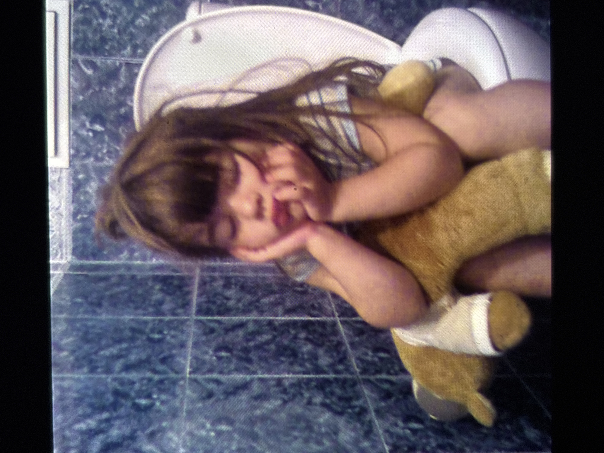 A young child, appearing to be between three and five years old, sits on a toilet seat. The child has dark hair and is wearing a striped t-shirt. They are holding a tan stuffed animal, which has a white bandage wrapped around one of its legs. The child's head is resting on their hands, and their eyes appear to be closed. The child is seated in a bathroom with blue tiled walls and a white toilet. The scene is not identifiable as being in Burgas, Bulgaria.