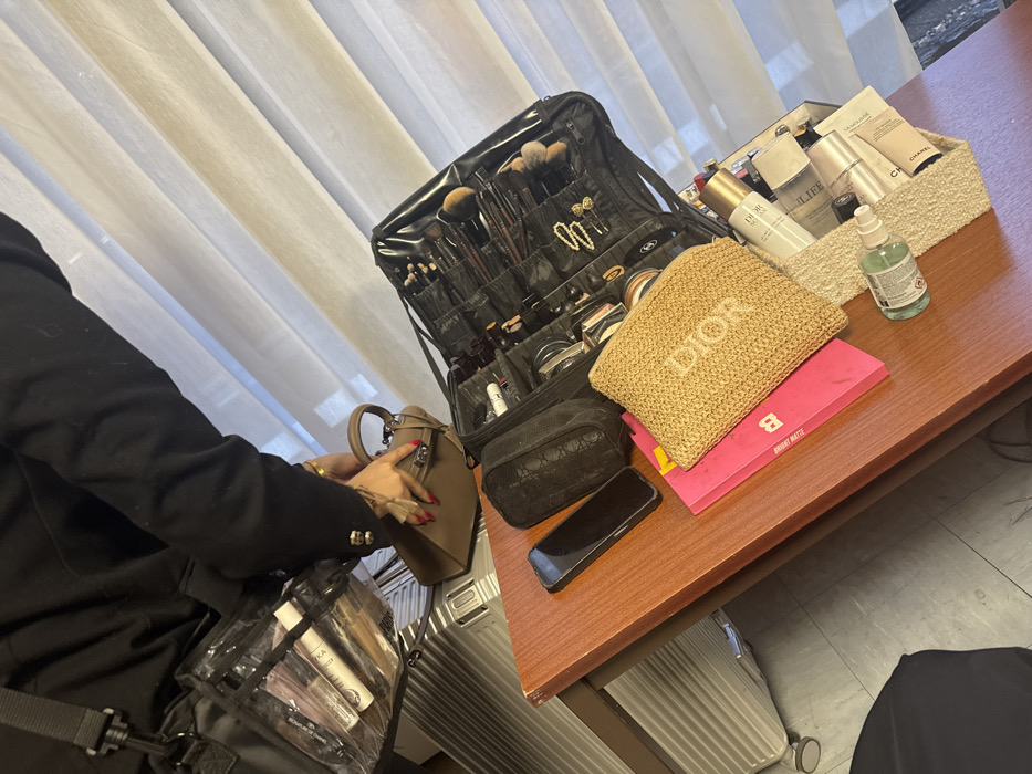 A makeup artist in a black blazer reaches for a taupe handbag next to a rolling luggage. The table is cluttered with a large black makeup kit, a tan Dior pouch atop a pink palette, a small black patterned pouch, and a smartphone. A basket filled with skincare products sits to the right. A bottle of hand sanitizer and additional luggage are also visible. The scene is lit by natural light filtering through sheer curtains.