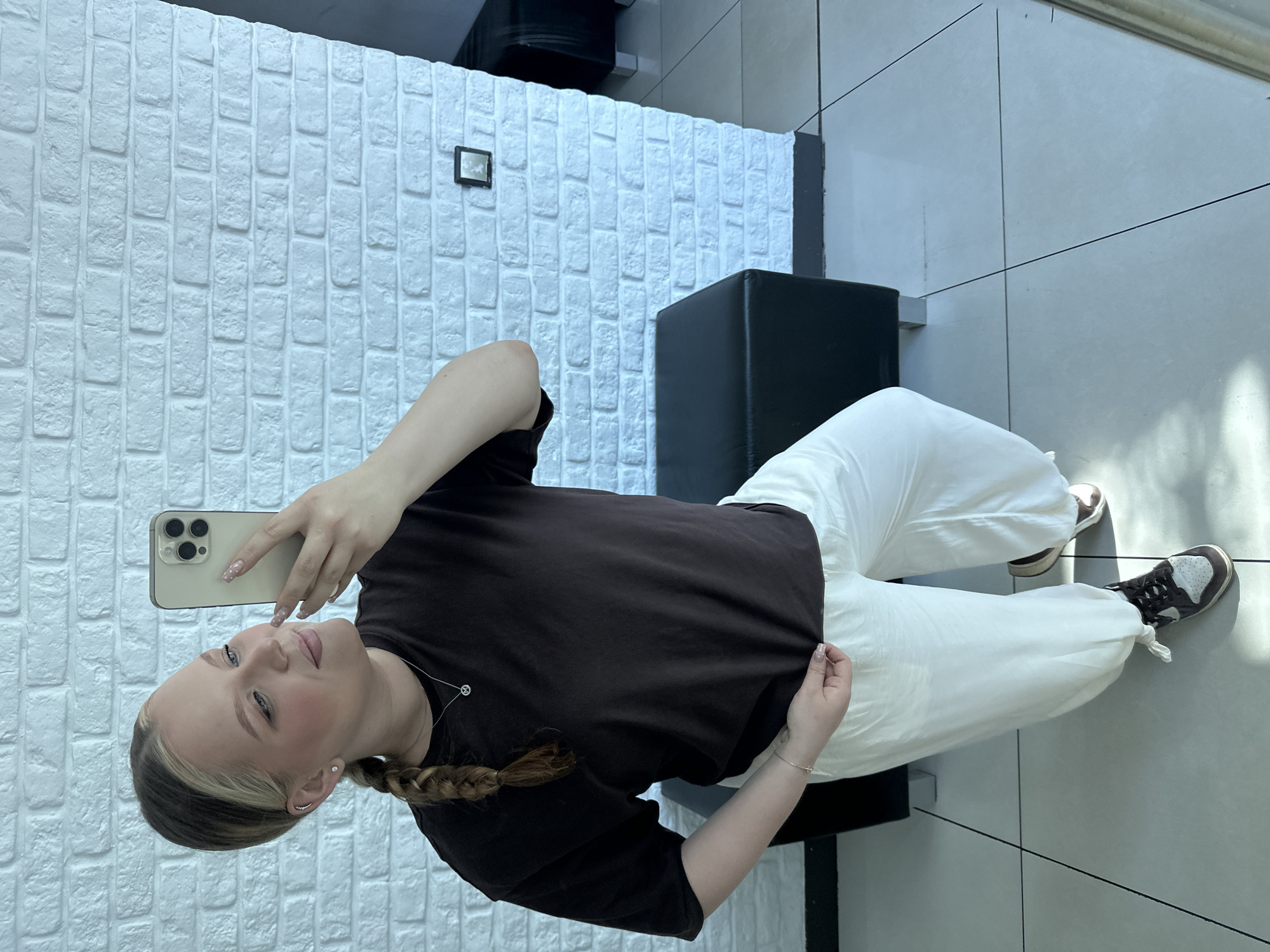 A woman is taking a selfie in a room with white brick walls and gray tile flooring. She is wearing a dark brown t-shirt, white pants, and brown and white sneakers. She has a blonde braid and is holding a gold smartphone. There is a black cube-shaped stool in the background. It is daytime. The location is Chaikovsky, Russia.