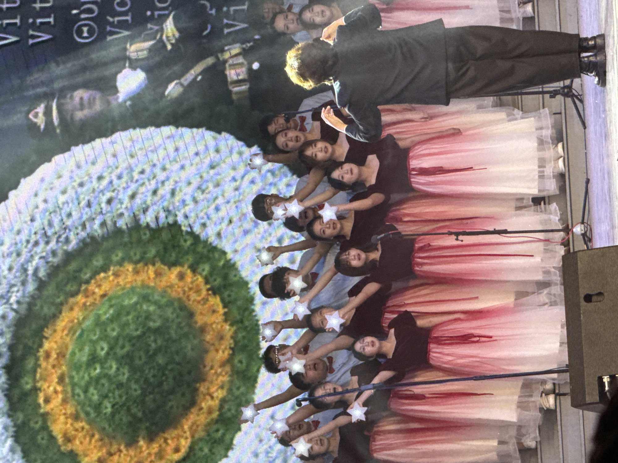 This image captures an indoor stage performance, likely a choral or musical event, in Guta, China. The scene features a group of young performers arranged in rows on a stage, backed by a large digital screen displaying a vivid, abstract circular pattern resembling a flower or target, with concentric rings of green, yellow, and blue hues.
The performers consist of both male and female individuals. The women wear black tops paired with elegant, layered skirts that exhibit a white-to-pink/red ombre effect. The male performers are dressed in lighter-colored shirts (possibly white or light blue) accessorized with bow ties. All performers are holding illuminated, star-shaped objects, extending them forward as a synchronized visual element of the act.
At the forefront, a conductor or leader, dressed in a dark suit, stands with their back to the camera, facing the performers and seemingly directing the ensemble. Microphones on stands are visible near the performers, confirming a vocal component to the performance. Stage equipment, including a dark speaker, is situated on the right side of the stage.
The background screen also projects images of other figures, including what appears to be a person in a military-style uniform. Fragments of text are visible on the screen, including repeated "Vit" and "Vict" possibly indicating "Victory," along with the Greek letter Theta (Θ). The setting suggests an evening event, with artificial lighting illuminating the stage and performers. The participants appear focused and engaged in their performance.