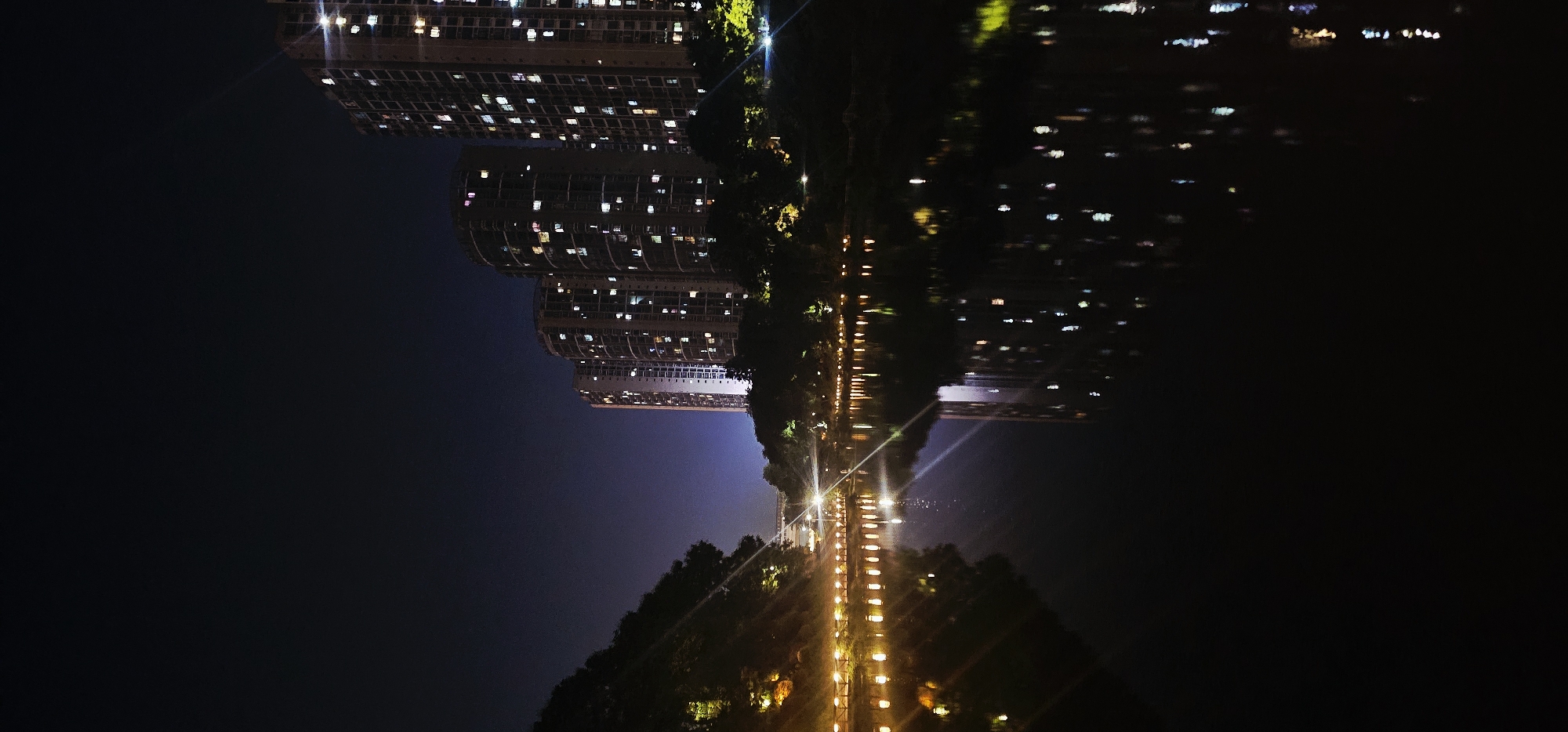 A night scene depicts a calm body of water reflecting the lights of a city. On the right, several tall, modern apartment buildings are illuminated with a multitude of windows glowing in various colors, including white, yellow, pink, and blue. These buildings are densely packed together. To the left of the buildings, a treeline separates them from the water.
Along the edge of the water, a pathway is lined with a series of evenly spaced, warm-toned lights, casting reflections on the water's surface. Further to the left, a hillside covered in dark foliage rises, also illuminated by some scattered lights. The sky above is a deep, dark blue, suggesting nighttime. The overall impression is one of urban development adjacent to a natural water feature, characteristic of areas like Bishan, China.