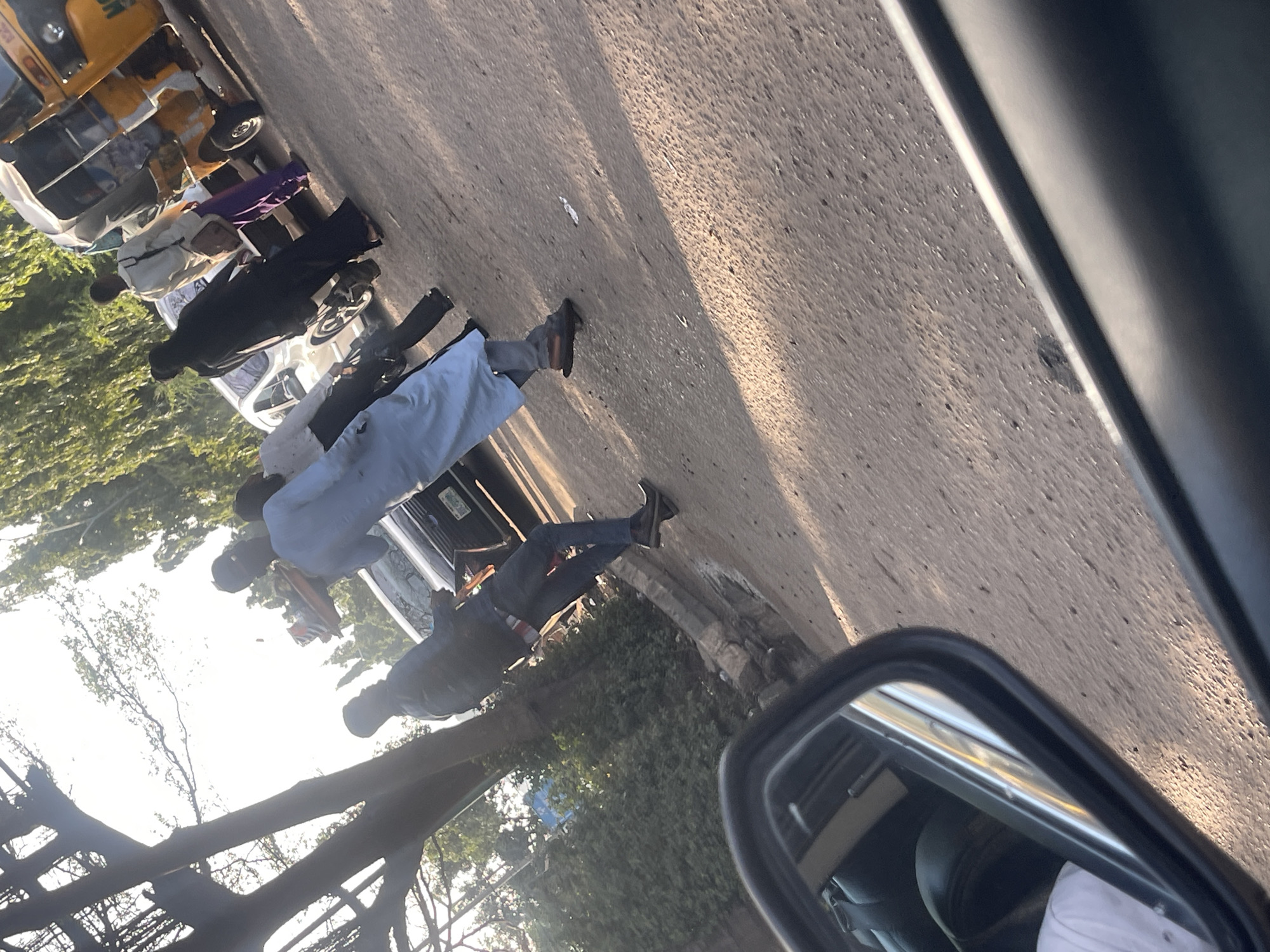 The image captures a street scene, presumably in Dutsen Alhaji, Nigeria, viewed from inside a car. A row of vehicles, including a yellow tricycle taxi, is parked along the side of the road. Several people are walking or standing along the street, some wearing traditional attire such as a light blue boubou. The setting appears to be a typical urban streetscape with trees and buildings visible in the background. The road surface is paved. A car's side mirror is prominent in the foreground.