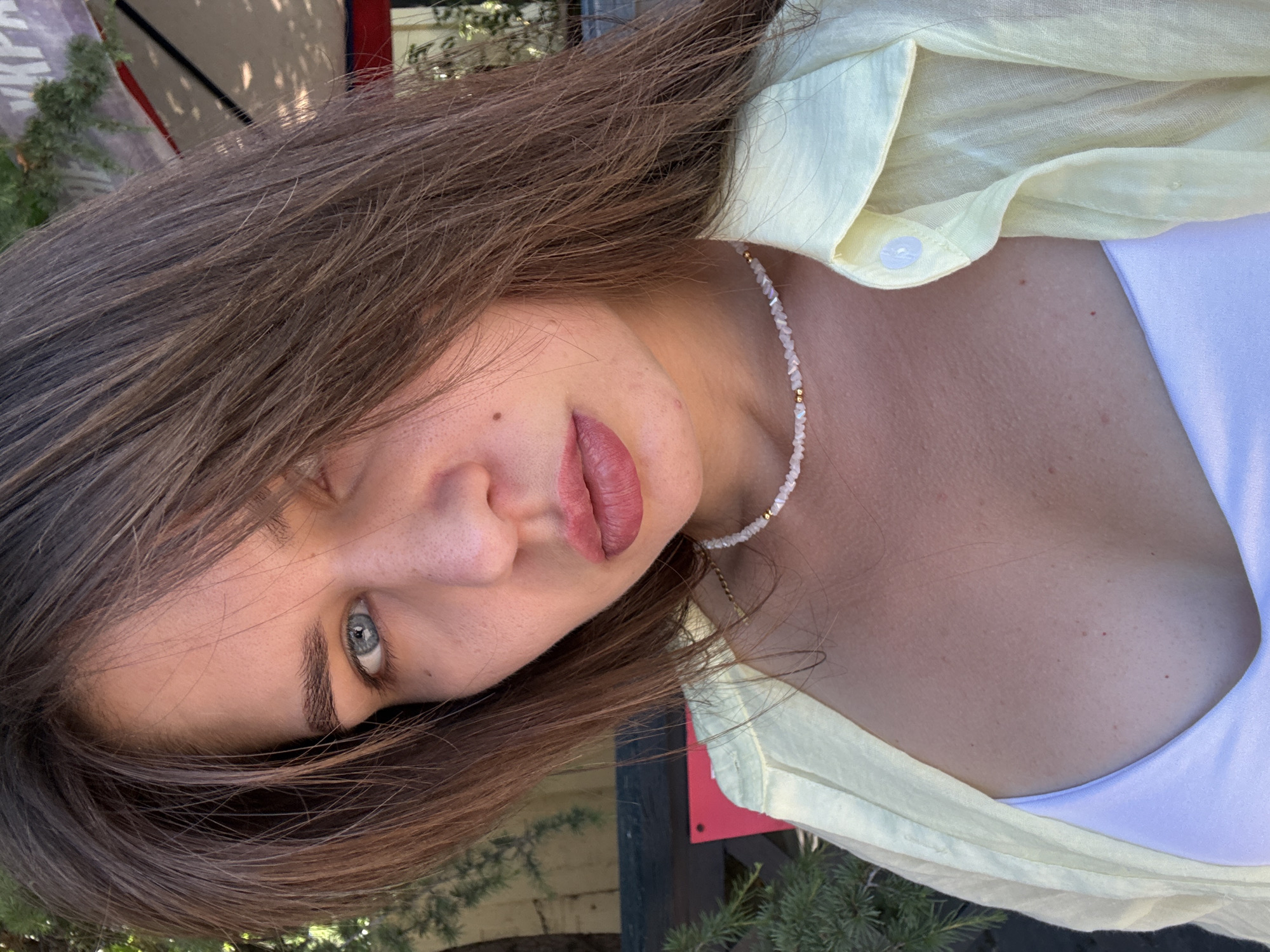 A medium shot captures a young woman with long, brown hair and blue eyes looking directly at the camera. She is wearing a light yellow collared shirt open at the neck, revealing a white top underneath and a delicate beaded necklace. The background is blurred, suggesting an outdoor setting with greenery and possibly a building with a red awning. The lighting indicates it is daytime, and the woman appears to be outside in a casual environment.