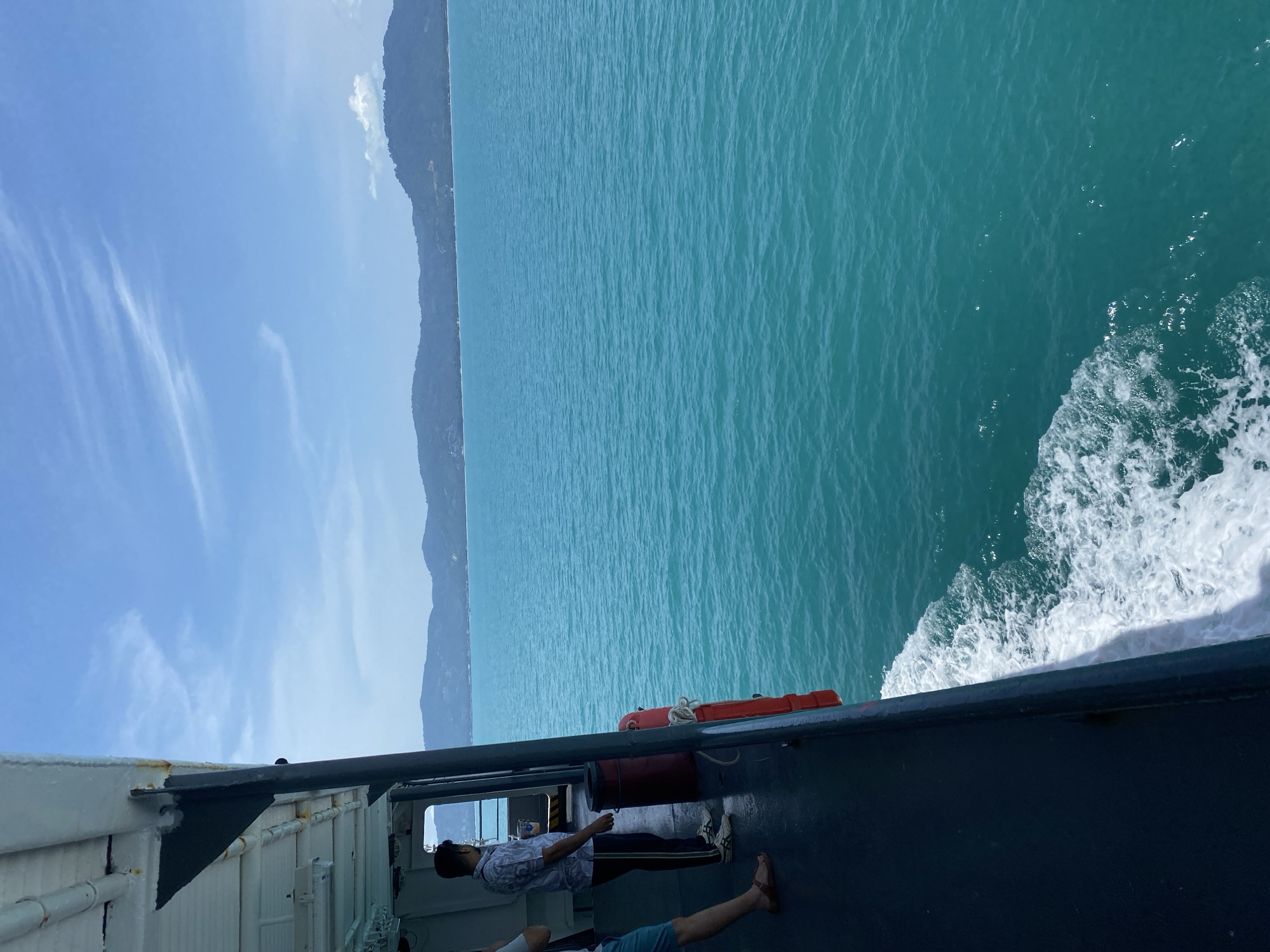 On a ferry crossing clear turquoise waters, a man in a patterned shirt and dark pants stands on the deck, looking out towards a distant, hazy mountain range. He holds a cup in his right hand. To his left, another person's legs are visible, wearing blue shorts and sandals. The ferry's dark blue hull curves along the left side of the frame, with white foam from its wake trailing in the water. A red life preserver is attached to the railing. The sky is a bright blue with scattered white clouds. The scene suggests a travel route between islands or along the coast in Thailand.