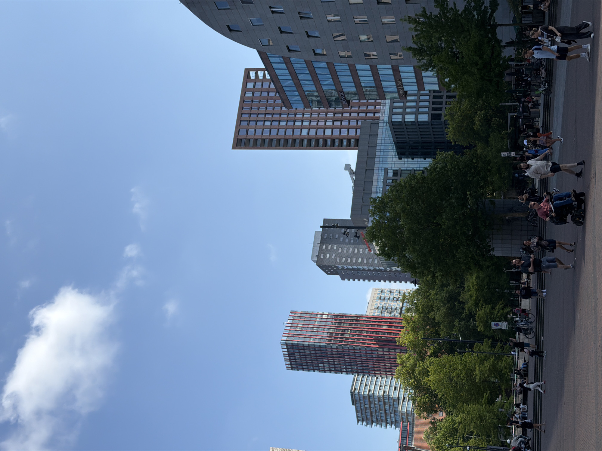 The image depicts a sunny day in Rotterdam, Netherlands, specifically showcasing the modern architecture of the city center. The foreground features a paved plaza where several people are walking, standing, and interacting. A person is seated in a mobility scooter, moving across the plaza. Other individuals are walking in groups or individually.
In the background, a cluster of tall buildings dominates the skyline. A prominent red and white striped skyscraper stands out, alongside other contemporary high-rises with glass facades and geometric designs. Lush green trees are interspersed between the buildings and in a park-like area in the middle ground, providing a contrast to the urban environment. The sky is a clear blue with a few scattered white clouds. The overall scene conveys a sense of urban life and modern development in Rotterdam.