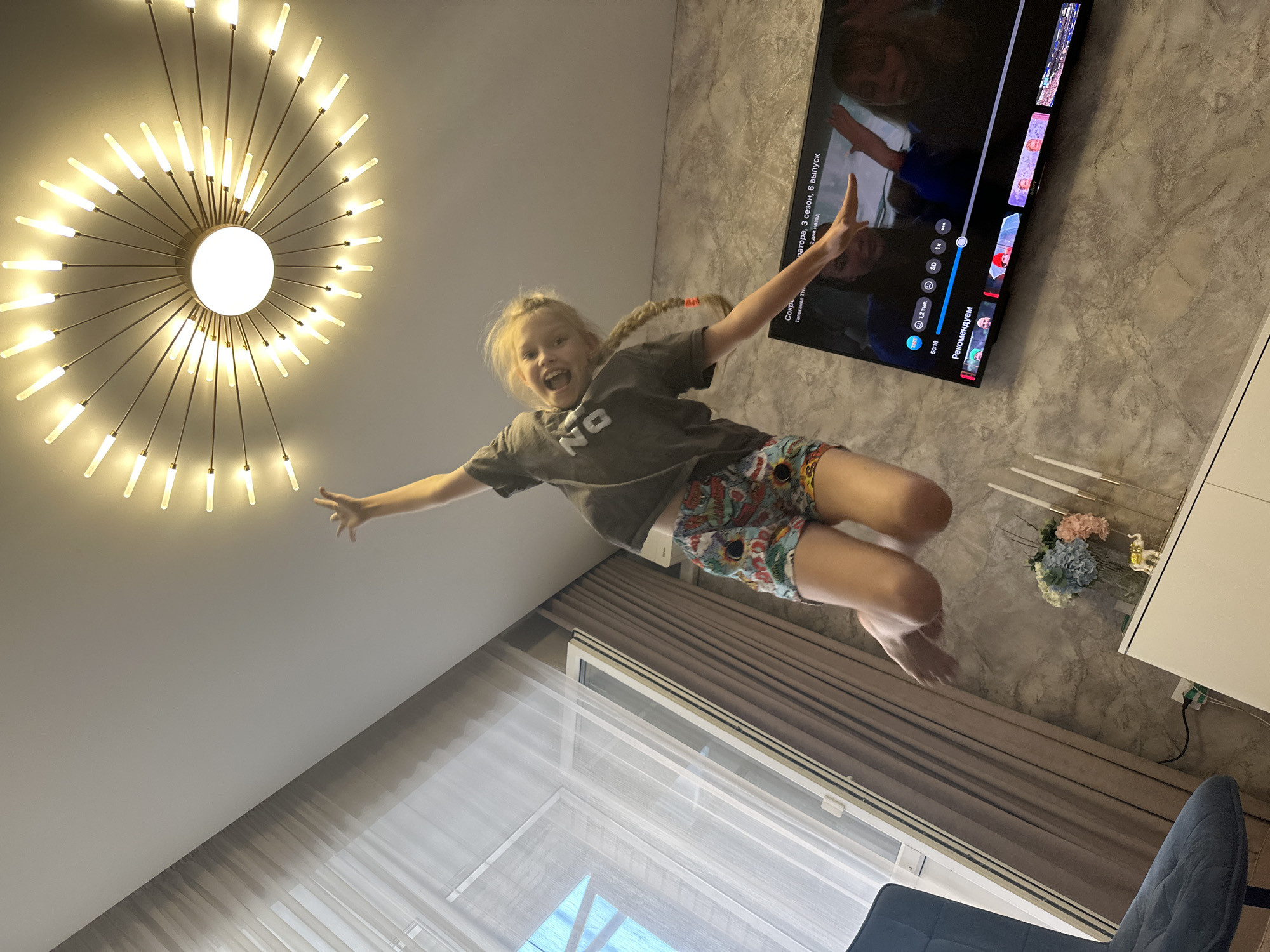 The image shows a young girl with blonde pigtails jumping in the air in what appears to be a living room. She is wearing a grey t-shirt with the word "NO" on it and colorful patterned shorts. Her mouth is open in a wide smile, and her arms are outstretched, suggesting joy and freedom.
Above her, a modern, starburst-shaped chandelier with many light bulbs illuminates the ceiling. To her right, a large flat-screen television is mounted on a textured grey wall. The TV displays a streaming service interface, showing episode information in Russian ("сезон, 6 выпуск" likely meaning "season, 6th episode") and a selection of recommended content.
The background includes floor-to-ceiling curtains, some sheer and some opaque, and a window. The lighting suggests it could be daytime, but the indoor setting makes it difficult to be certain. The overall atmosphere is playful and energetic, centered around the girl's enthusiastic jump. The presence of the TV and home decor indicates a domestic setting.