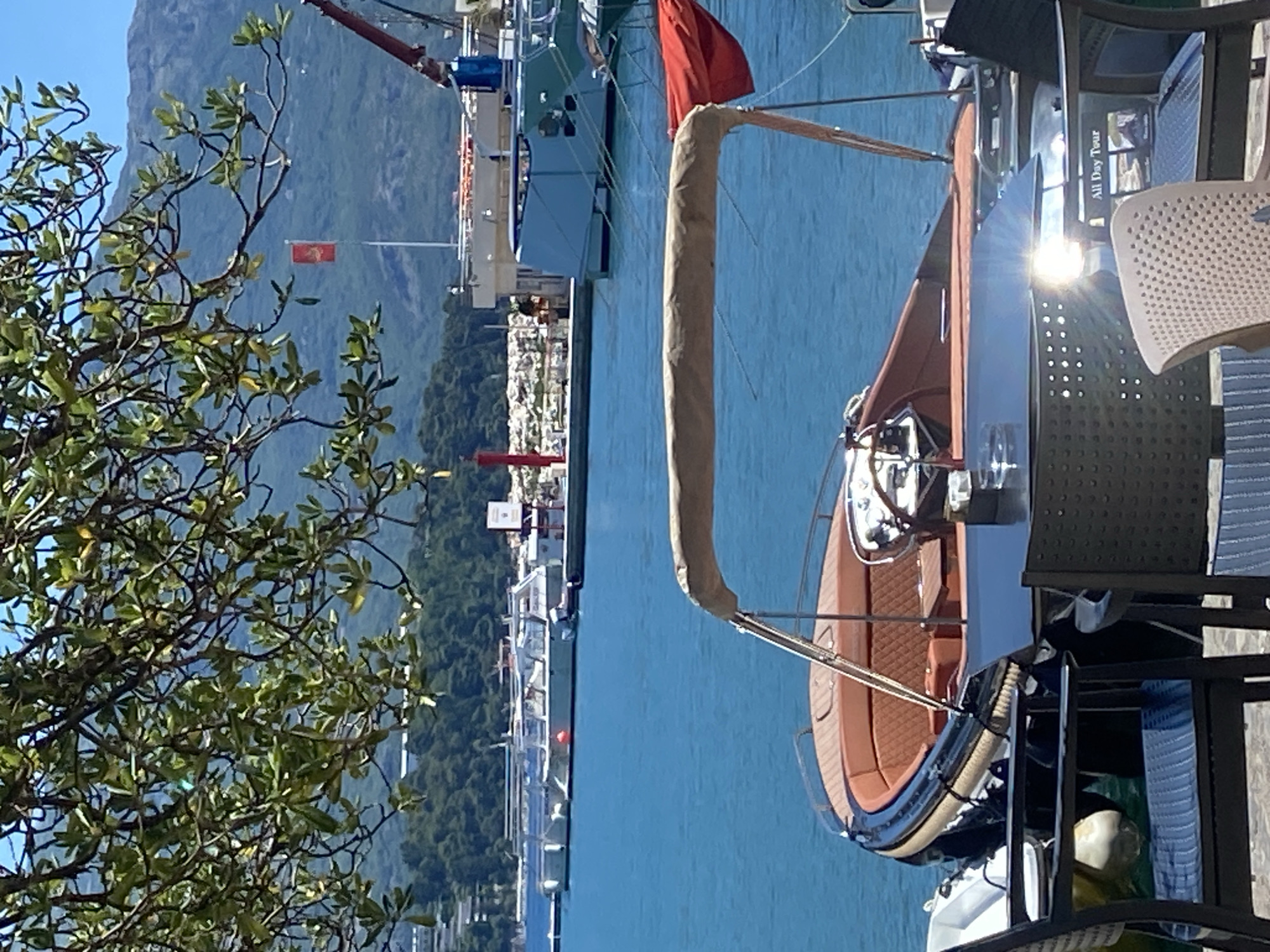 In Budva, Montenegro, a luxury motorboat with a tan canopy and brown leather seating is docked near a table set for outdoor dining. The boat is partially obscured by a table and chairs in the foreground. Beyond the boat, the clear blue water of the Adriatic Sea is visible, with other vessels and a marina in the background. A lush green hillside and distant mountains form the backdrop. The scene is bathed in bright sunlight, casting a lens flare on the table's surface. A red flag, possibly the flag of Montenegro, is visible flying from a pole in the distance. The overall impression is one of a serene, upscale waterfront setting.