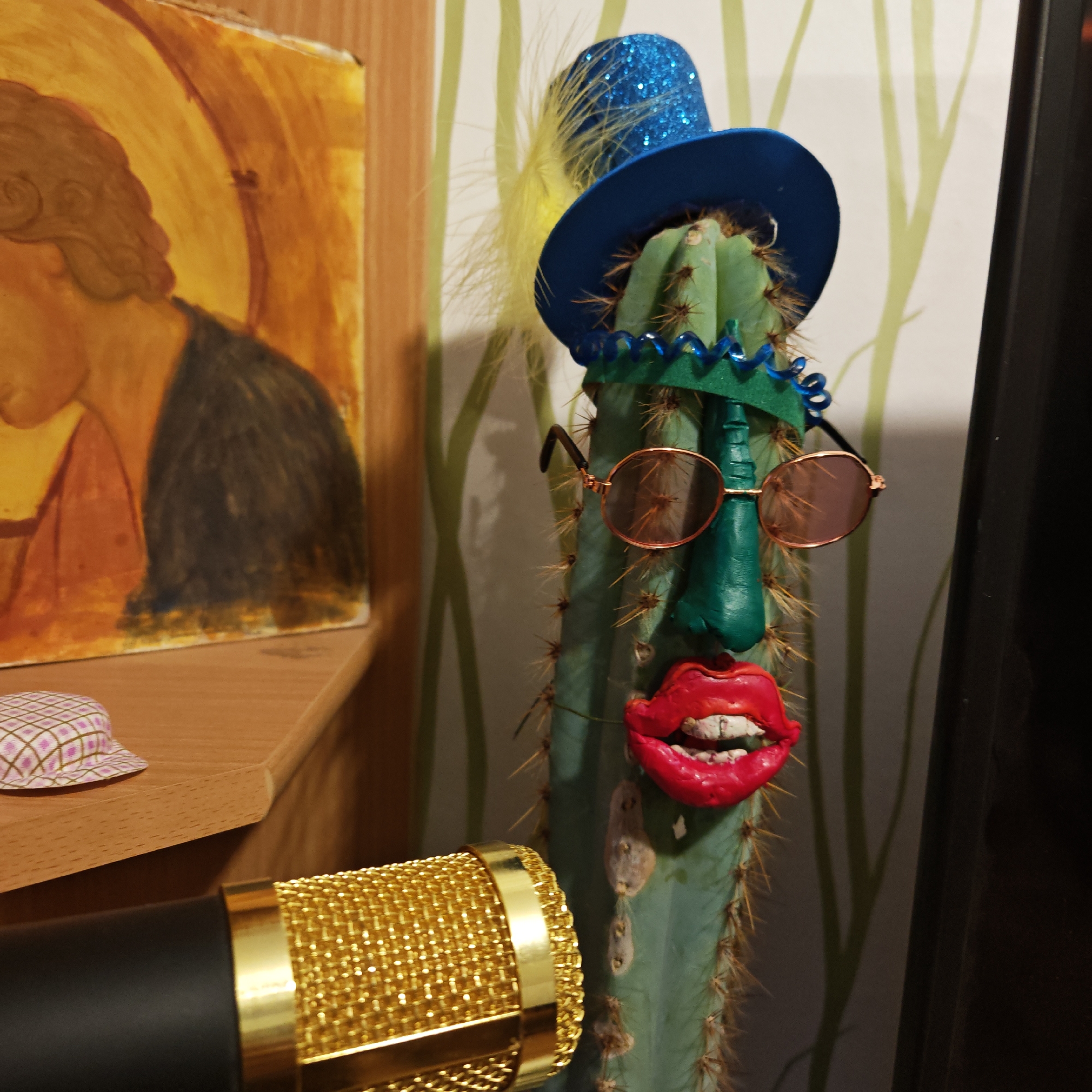 A medium shot depicts a decorated cactus, resembling a character, positioned in front of a wall with light green, stylized tree branches. The cactus is adorned with a blue glittery top hat with a yellow feather, a blue beaded garland around its "neck," rose-tinted sunglasses, a green nose, and large red lips with exposed teeth. A gold microphone is situated to the left in the foreground. Behind the cactus and to the left is a wooden surface with a painting of a person's head and torso, possibly an icon. A small, patterned hat sits on the wooden surface.
There is no reliable information in the image to confirm the location is Rostov, Russia.