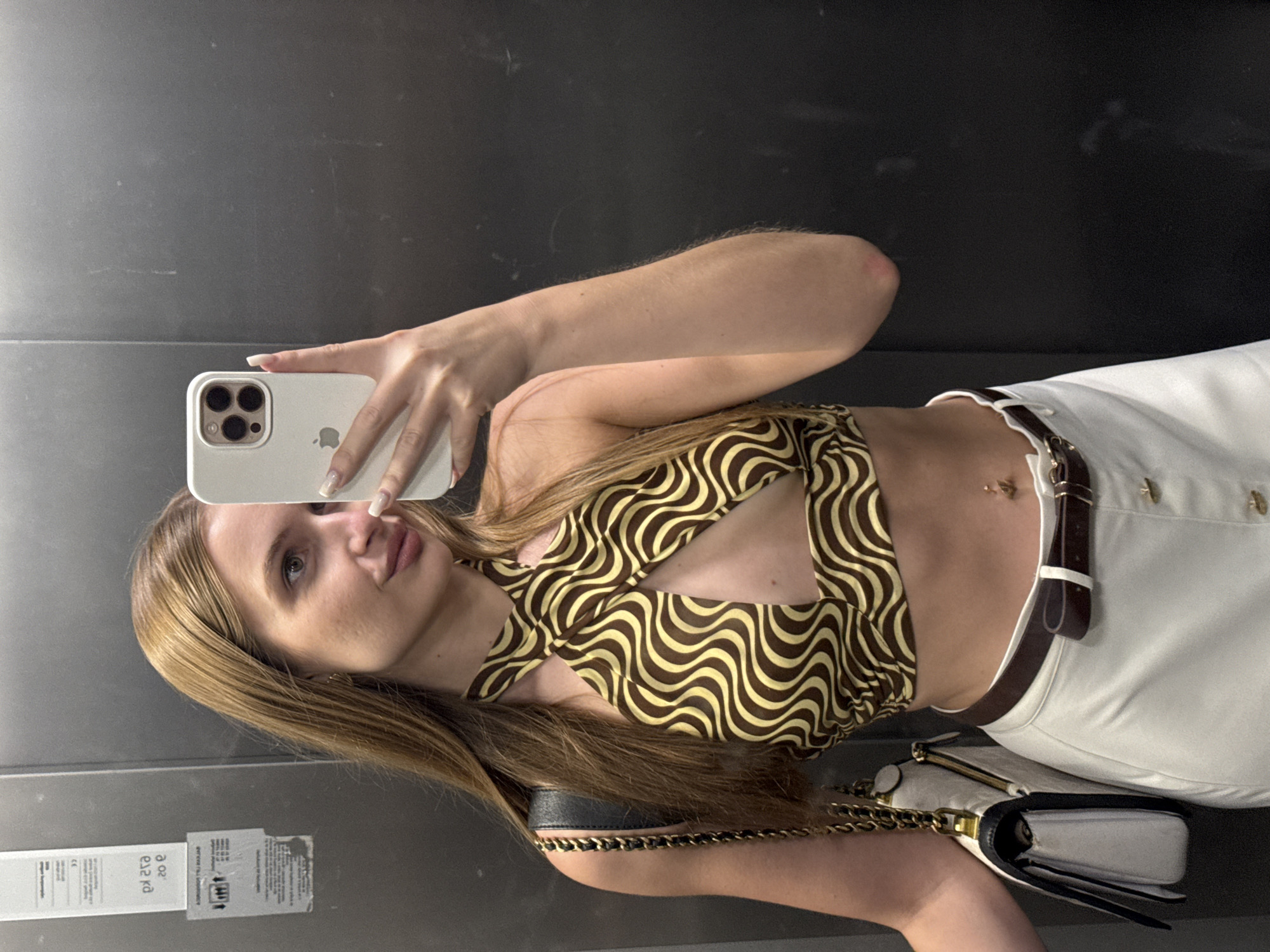 A young adult female is depicted taking a mirror selfie inside an elevator, presumed to be in Budva, Montenegro. She has long, straight, light brown hair and is looking towards her reflection. Her right hand holds a white iPhone, clearly showing its triple camera setup and Apple logo, partially obscuring her face. She wears a halter-neck crop top featuring a brown and light yellow wavy pattern with a keyhole cutout at the sternum. Below, she wears high-waisted white trousers or a skirt with visible buttons. A dark brown leather belt with a white loop cinches her waist, and a gold-colored navel piercing, appearing as the number "4", is visible on her abdomen. A white shoulder bag with black details and a gold chain strap hangs on her left shoulder. The elevator interior features dark grey, possibly metallic, walls with visible scratches and smudges, and a partially reflected sign with indistinct text on the left.