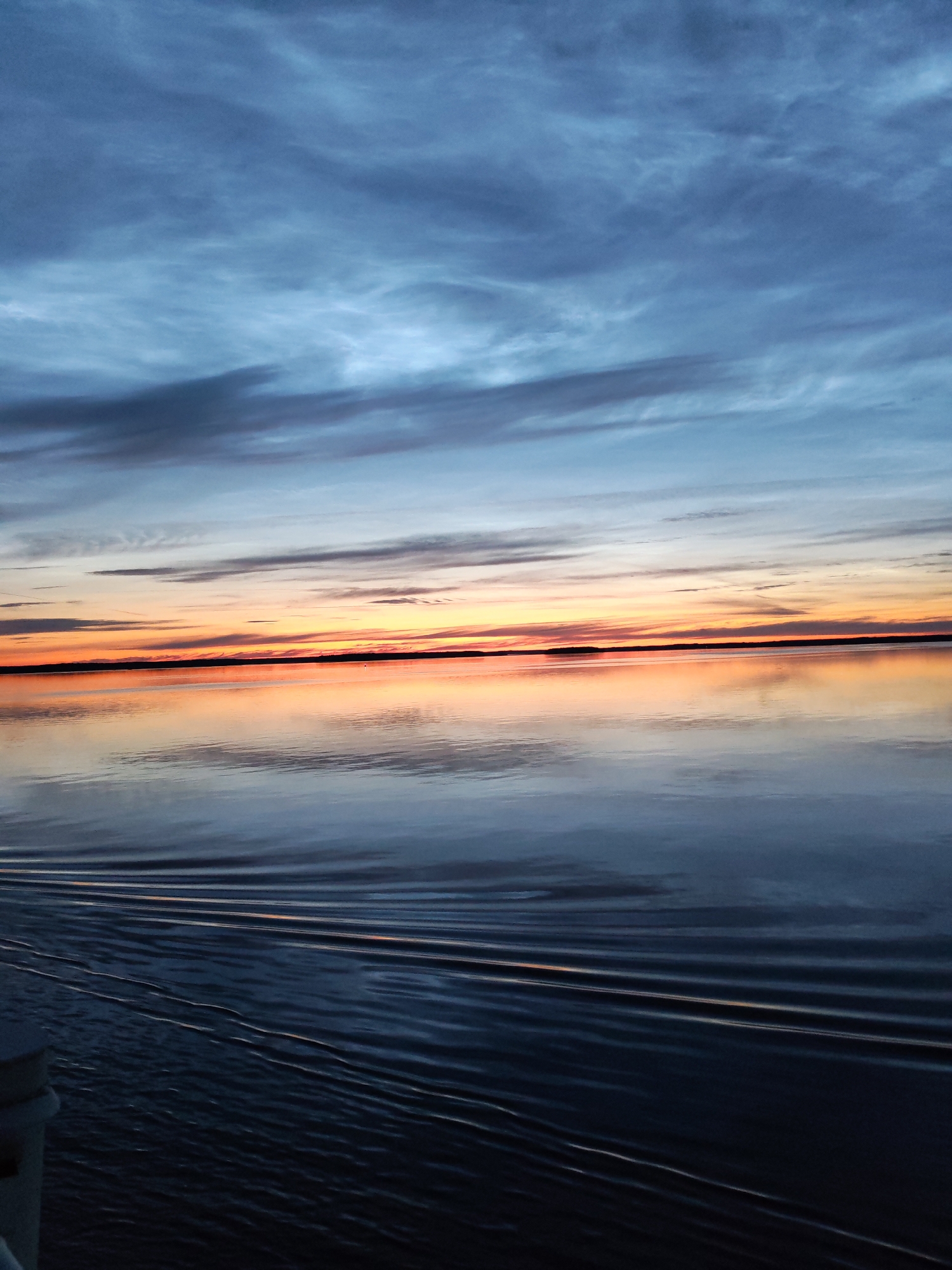 The image depicts a serene sunset over the Sheksna River in Sheksna, Russia. The sky is a gradient of colors, transitioning from deep blue and gray clouds at the top to vibrant oranges and yellows near the horizon. The calm water perfectly reflects the sky's colors, creating a mirrored effect. Gentle ripples on the water's surface suggest movement, possibly from a boat or a slight breeze. In the foreground, a portion of a white railing or structure is visible, indicating the vantage point is from a vessel or pier. No people are visible in the scene, which is dominated by the natural beauty of the sky and water.
