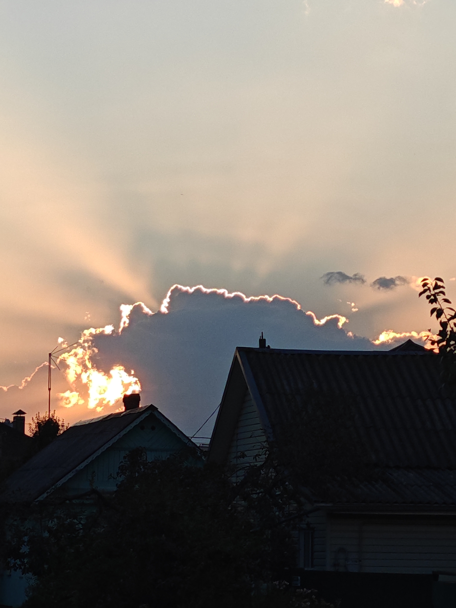The image captures a dramatic sunset over a residential area in Homyel, Belarus. Silhouetted against the warm, orange and pink hues of the sky are the rooftops of several houses. Prominent in the frame is a large, dark cloud with its edges illuminated by the setting sun, creating a halo effect. Rays of light emanate from behind the cloud, piercing through the sky in a crepuscular display. In the foreground, the dark shapes of trees and house structures are visible, further emphasizing the silhouette effect. The overall scene evokes a sense of natural beauty and tranquility as day transitions into night.