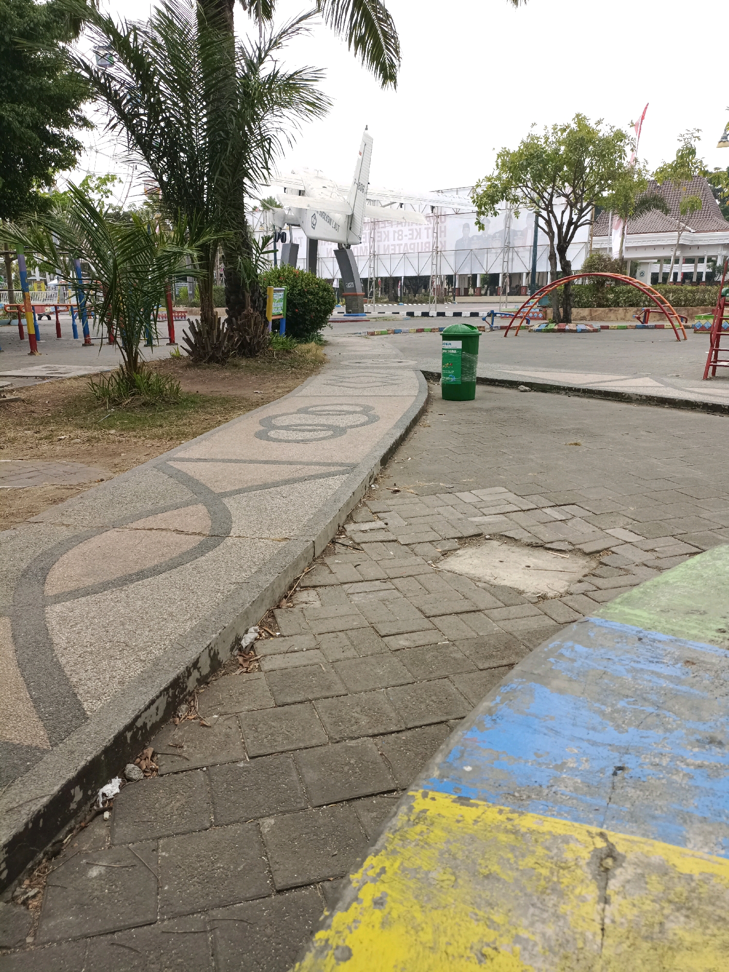 The image depicts an outdoor scene in the city of Lamongan, Indonesia. In the foreground, a paved walkway with a decorative pattern curves to the left. The walkway is bordered by a concrete curb and a patch of damaged brick paving. A green trash can is positioned on the right side of the walkway. In the middle ground, a playground area with colorful equipment is visible, including red and yellow arched structures. A palm tree stands to the left of the walkway, and a large structure resembling an airplane is displayed prominently in the background. The background also shows buildings and trees. The scene appears to be a public park or recreational area.