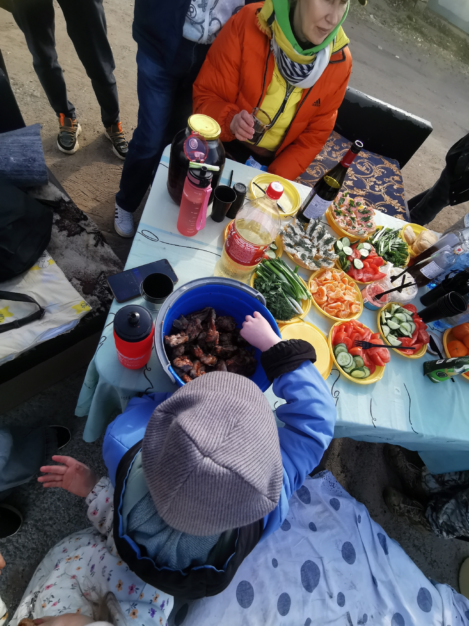 An outdoor gathering is captured from an overhead perspective, showcasing a picnic setup. Several individuals are present, including adults and children. On the left, a person's lower body is visible, wearing dark pants and sneakers. Another adult, dressed in a bright orange and yellow padded jacket with an Adidas logo and a green and white scarf, is seated, holding a glass. A child in a blue jacket and grey knitted hat is reaching into a large blue bucket filled with cooked meat, possibly shashlik or grilled kebabs. Part of another child's head and a floral patterned dress are visible in the foreground.
The central focus is a light blue tablecloth laid over a table, laden with a variety of food and drinks. Numerous yellow plastic bowls are filled with fresh produce: sliced cucumbers, chopped tomatoes, green onions, dill, and peeled mandarin segments. Other bowls contain spreads or salads, one with a pinkish mixture and another with a white, dill-garnished dish. Bread rolls are also on the table.
Beverages include a large glass jar with a dark liquid, a pink water bottle, a red-labeled clear plastic bottle containing a yellow liquid (resembling a repurposed Coca-Cola bottle), a red wine bottle (partially visible text "CRIV..."), and a red thermos. Black plastic cups and cutlery are scattered among the dishes. A smartphone lies on the table next to a black mug. The ground beneath the table is bare earth, suggesting a casual outdoor setting. The bright lighting indicates daytime. People are dressed in casual, warm clothing, suitable for a cooler outdoor environment. A white blanket with large dark polka dots is partially visible on the ground.