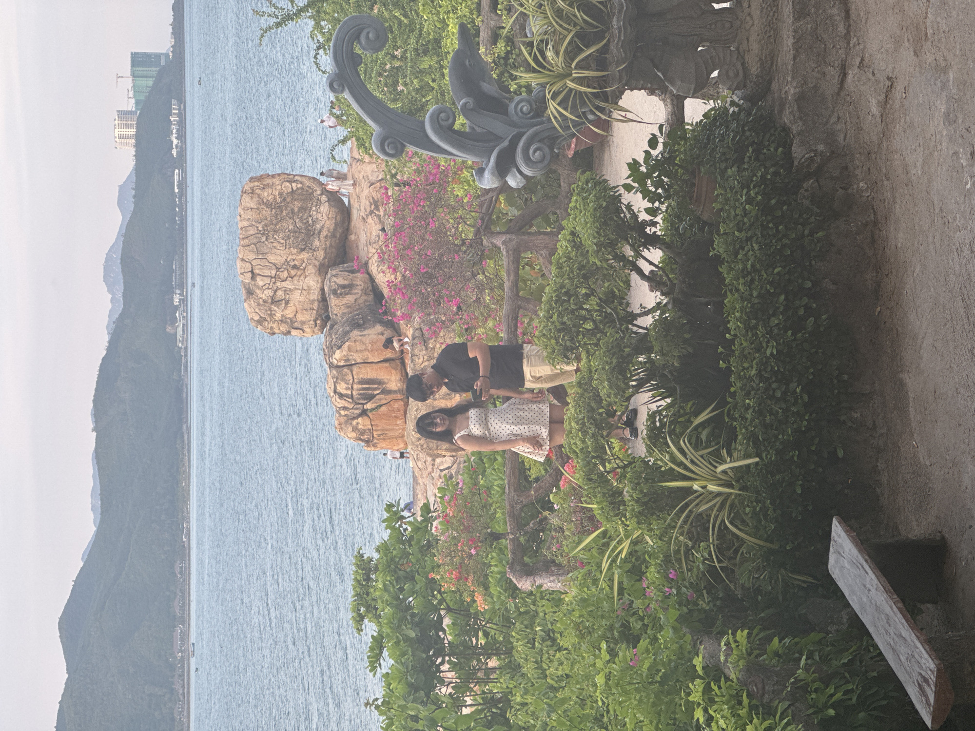 This image depicts a coastal scene in Bac Nha Trang Ward, Vietnam. In the foreground, lush green foliage surrounds a pathway and a rustic wooden bench. A dark gray sculpture of a dolphin emerging from waves is prominently placed on the right. In the mid-ground, a young couple stands near a weathered wooden fence, with the woman in a white polka dot dress and the man in a black t-shirt and khaki shorts, both looking at a mobile device. Behind them, large, weathered rock formations are visible, with a vibrant pink flowering bush adding a splash of color. In the background, the calm, blue sea stretches towards a hazy mountain range, with a few boats dotting the water and buildings visible on the distant shore. The sky is overcast, suggesting a mild climate.