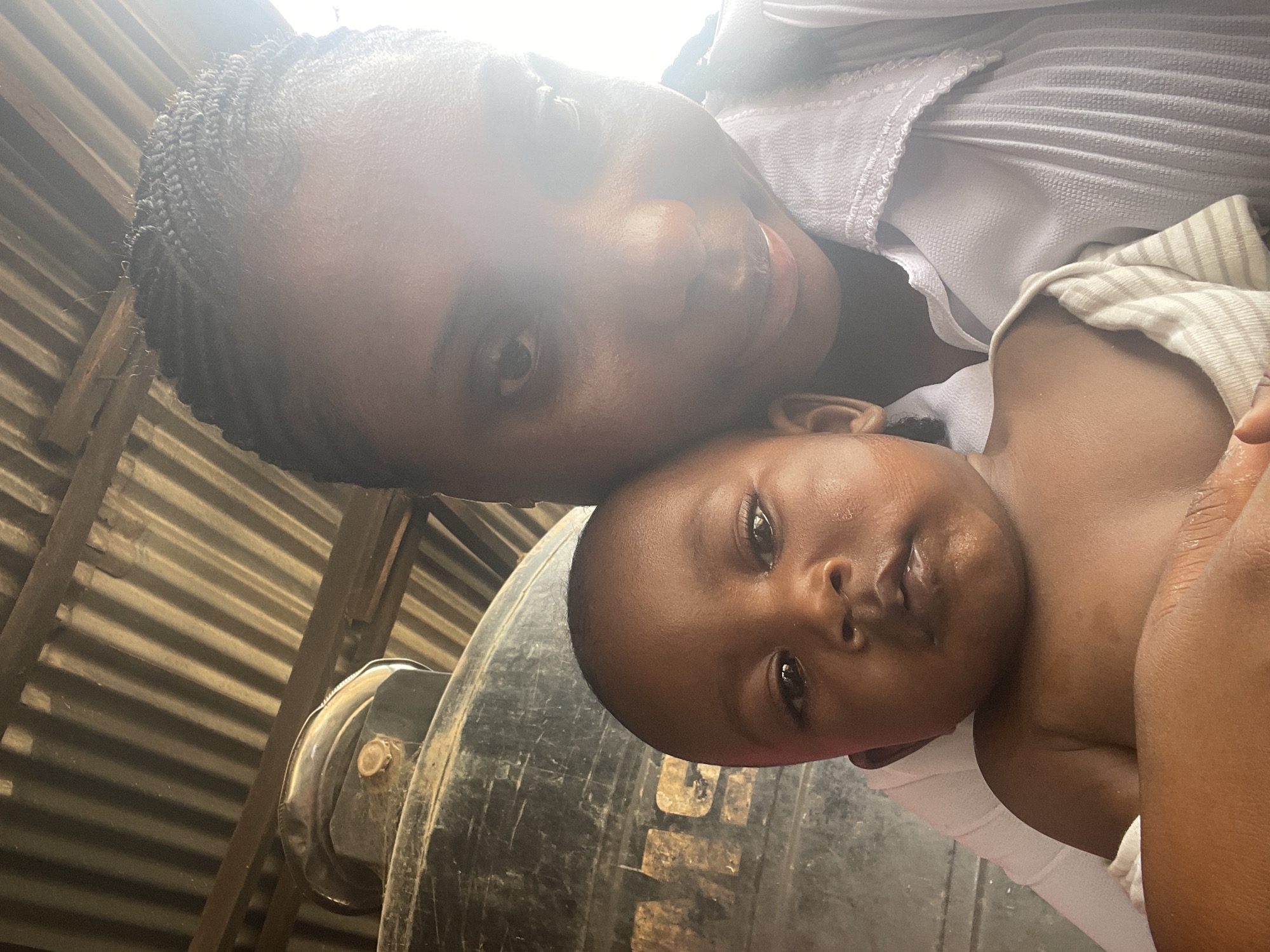 A woman with dark skin and braided hair is holding a baby. The baby also has dark skin. They appear to be in an outdoor setting, possibly near or under a corrugated metal structure. A large, black tank is visible in the background. The photograph appears to have been taken at an upward angle. The scene is assumed to be in Jos, Nigeria.