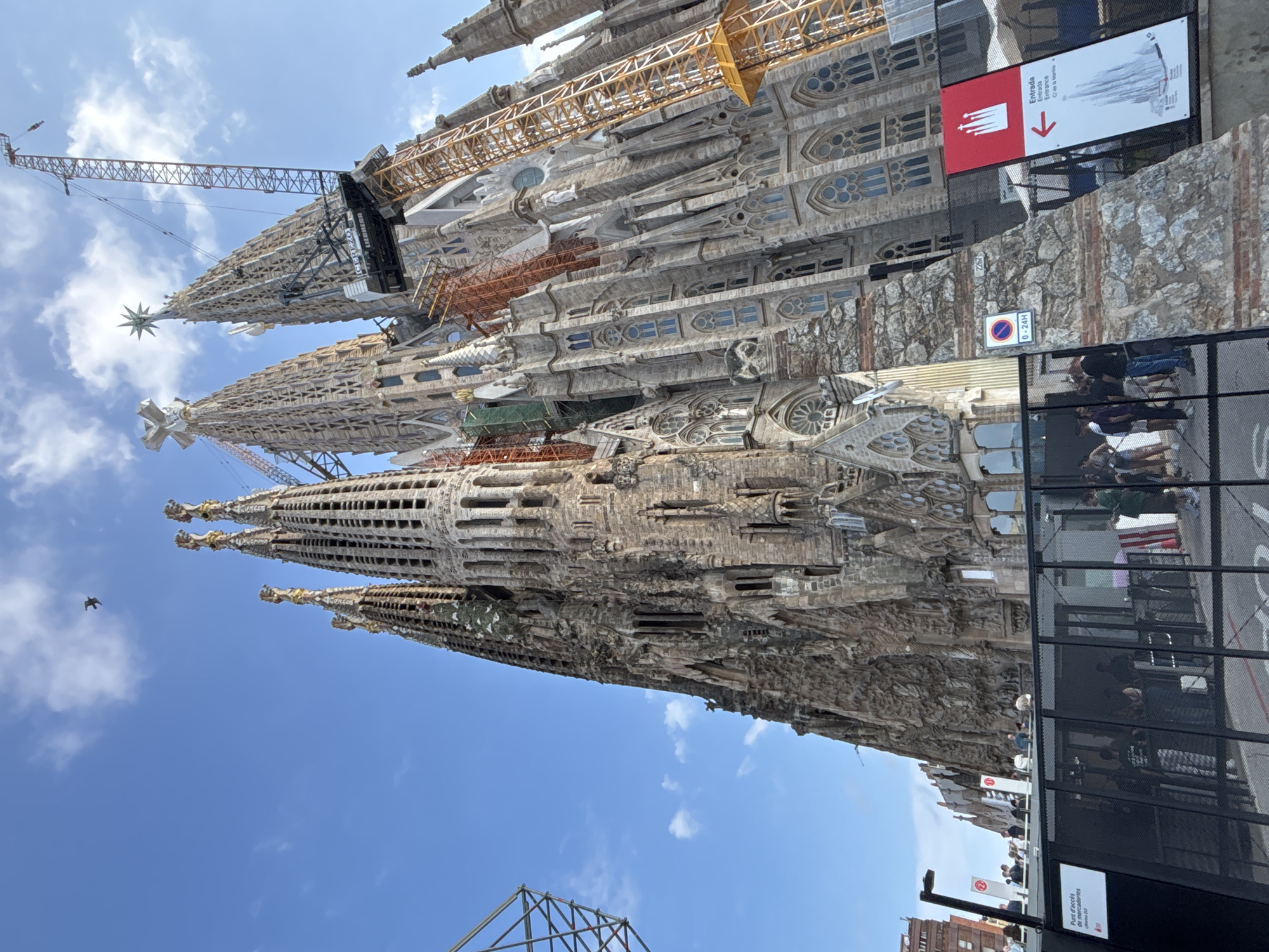 This image captures a majestic view of the Sagrada Familia basilica in Barcelona, Spain, under a bright blue sky with scattered white clouds, indicating daytime. The colossal Gothic and Art Nouveau structure dominates the frame, showcasing its intricate stone facade, ornate spires, and detailed carvings.
Several large construction cranes and scaffolding, visible in yellow and orange, are actively engaged around the upper parts of the basilica, highlighting the ongoing construction of this iconic landmark. One prominent spire is topped with a luminous star, while another features a cross-like finial.
In the foreground, a black metal fence separates the viewer from the basilica. Behind this fence, several individuals can be seen, appearing to be visitors or staff. Various signs are attached to the fence and a stone wall. A red and white sign with a downward arrow indicates an "Entrada" (Entrance), displaying the text: "Entrada Entrada Entrance C/ de la Marina" and "Anf". Another sign on the fence reads "Punt d'accés de mercaderies" (Goods access point). A circular sign displays "0-24H", likely denoting specific operating or access times. The number "2267" is also visible on the stone wall.
A small bird is captured in flight near one of the towering spires, adding a sense of scale to the grand architecture. A partial metal framework, possibly related to construction, is visible in the bottom left corner. The image conveys the impressive scale and the continuous, decades-long endeavor of completing Antoni Gaudí's masterpiece.