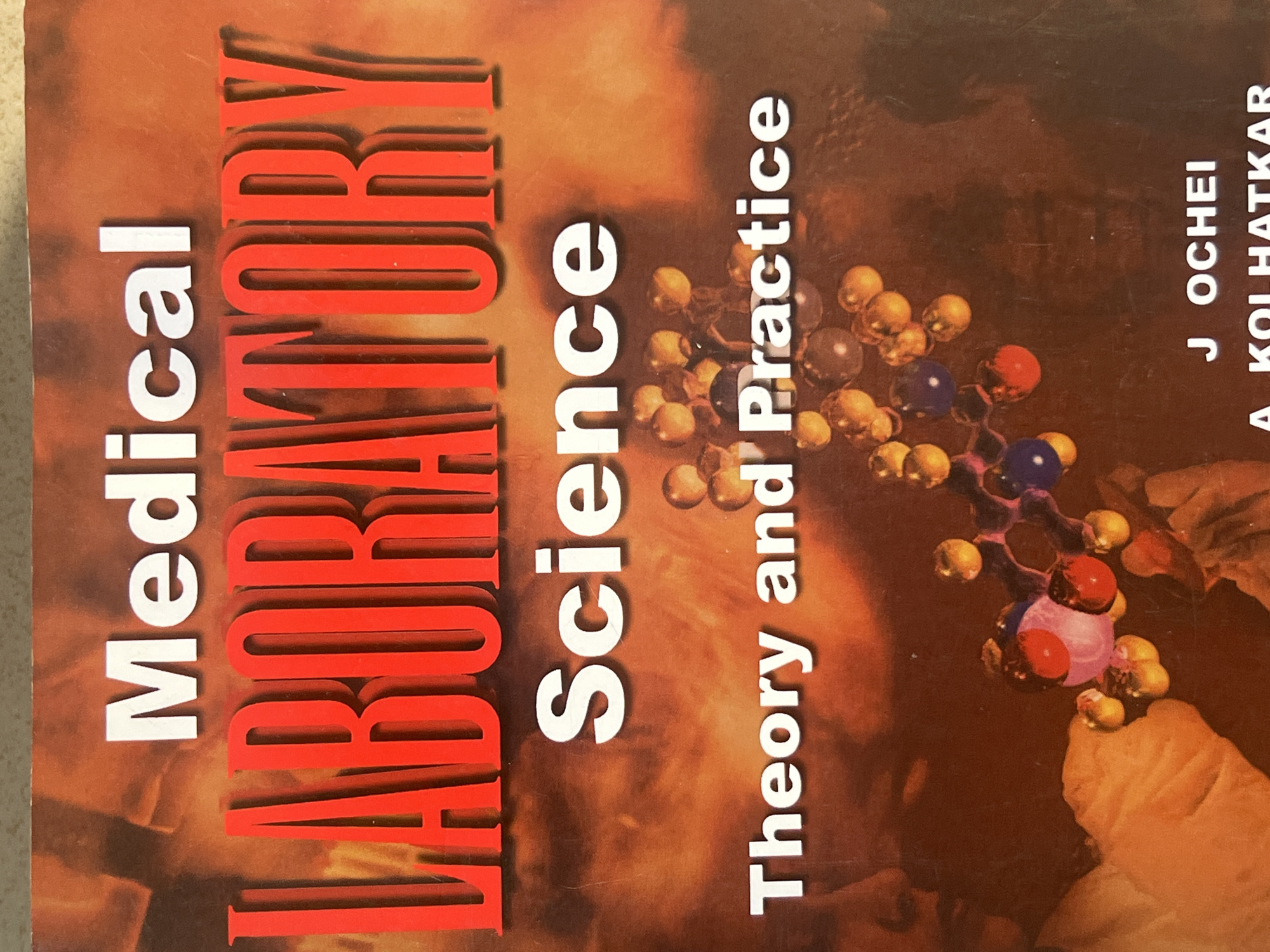 This is a close-up image of the cover of a book titled "Medical LABORATORY Science Theory and Practice". The title is presented in large, bold lettering, with "Medical" and "Science" in white, and "LABORATORY" in red. Below the title is a molecular diagram of a chemical structure, depicted with colorful spheres and connecting lines. To the left of the molecular diagram, a gloved hand is visible, holding what appears to be a beaker or a test tube with a pink substance inside. In the bottom right corner of the cover, the names "J OCHEI" and "A KOLHATKAR" are printed, likely the authors of the book. The background of the book cover is a blurred image with warm, reddish-orange tones, suggestive of flames or an abstract fiery design. There is no indication of any specific location within the city of Jauro Yinu, Nigeria, or any urban environmental elements present in the image.
