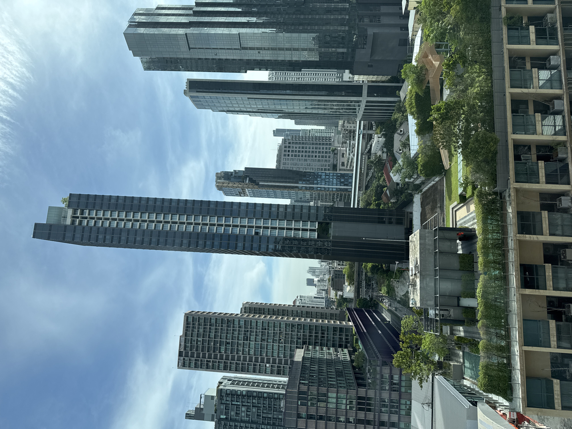 The image is an eye-level perspective of the urban landscape in Bangkok, Thailand, composed mainly of skyscrapers and greenery, and shot during the day. The sky appears light blue with some wispy clouds. The dominant features are the tall modern buildings with glass facades that stretch toward the sky. Among these buildings is the King Power MahaNakhon skyscraper. There is a rail system cutting through the landscape. The buildings are interspersed with trees and garden environments. Overall, the scene conveys a sense of urban density.
