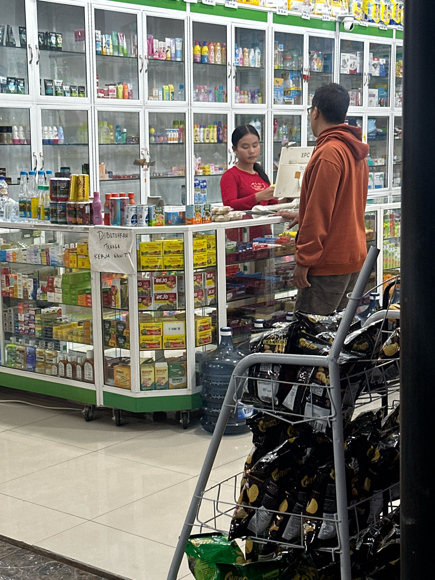 The image shows the interior of a shop, likely a convenience store or a small supermarket, located in the City of Binjai, Indonesia. The shop is filled with shelves stocked with various products, including food items, toiletries, and household goods. The shelves are illuminated and organized, with many items displayed in clear plastic packaging. In the foreground, a man wearing an orange hoodie and shorts is standing and looking towards the counter. A young woman, wearing a red long-sleeved shirt, is behind the counter, appearing to be interacting with the man. A sign written in Indonesian, which translates to "Looking for female workers," is displayed on a shelf near the counter. Several large bottles of water are visible, as are bags of chips on a display rack. The floor is tiled, and the overall impression is that of a bustling retail environment.