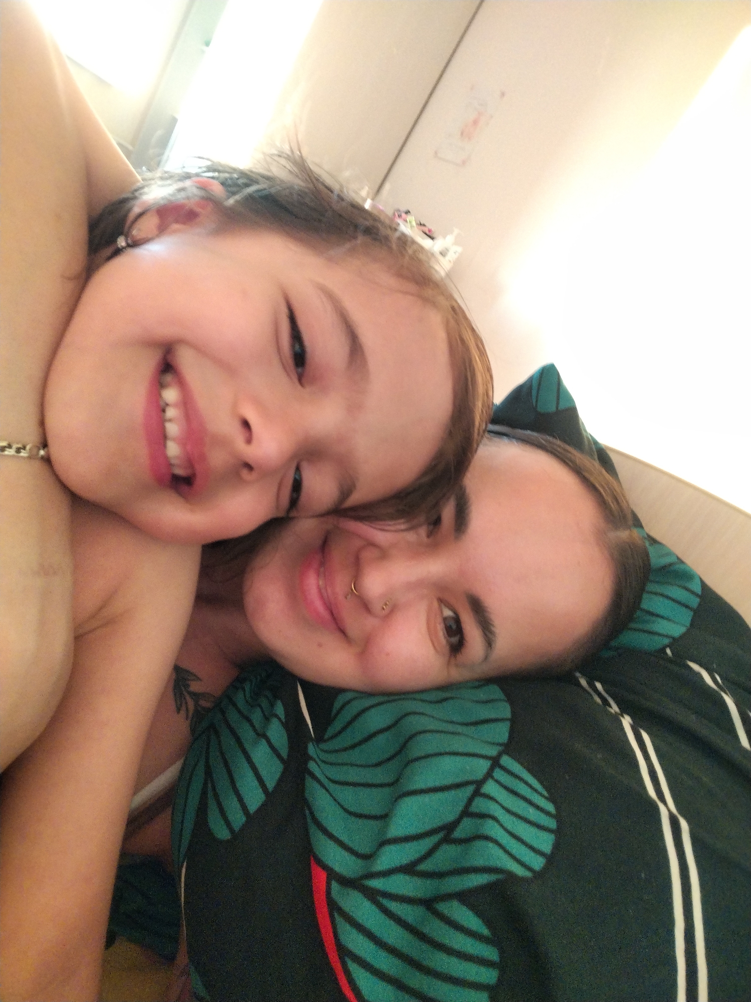 A close-up, candid shot depicts a young girl and an adult woman in an intimate embrace. The girl is smiling broadly, displaying her teeth, and appears to be resting her head on the woman. The woman, also smiling, has a nose piercing and is lying on a pillow with a green and black floral pattern. A tattoo is visible on the woman's shoulder. The scene is illuminated by a bright, presumably indoor light source. The location and the city of Noviy, Russia is not discernable from the image.