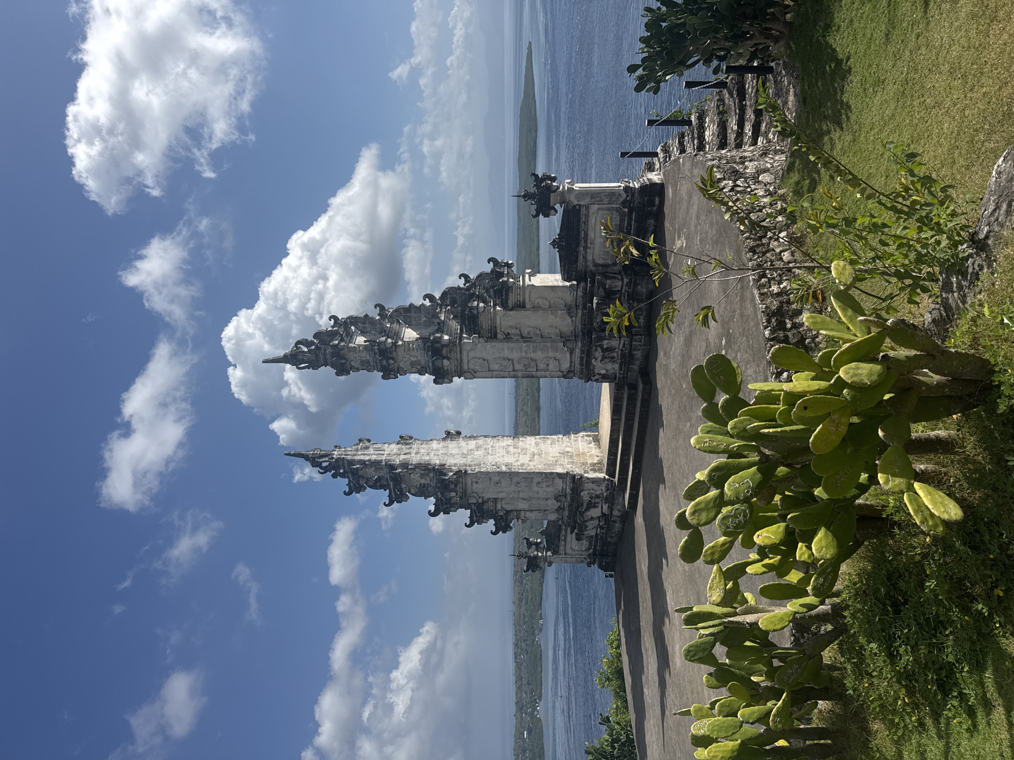 A stone temple entrance with ornate carvings stands on a cliff overlooking the ocean in Nusa Penida, Indonesia. The temple is flanked by two tall spires, and a wide stone pathway leads up to it. In the foreground, prickly pear cacti grow in a grassy area. The sky is a vibrant blue, dotted with fluffy white clouds. The ocean is calm and a deep blue, with a green island visible in the distance.