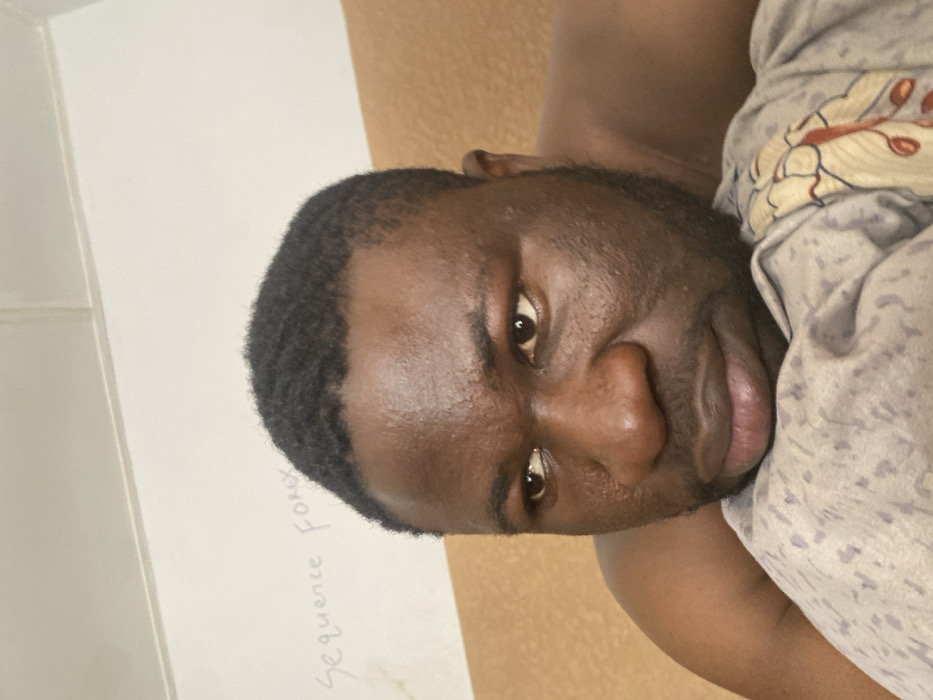 An adult male with dark skin, short dark wavy hair, and a short beard is depicted in an indoor setting within Udu, Nigeria. He is positioned with his head or upper body resting on a light grey patterned fabric, possibly a pillow or blanket, which features a faint floral or leaf design in muted colors. The man's bare right shoulder is visible, suggesting he is shirtless or partially clothed. He looks towards the left side of the frame with a neutral or contemplative expression. In the upper left background, a light-colored wall is visible, on which the handwritten text "Sequence Forex" is faintly discernible. A solid light brown or beige wall surface is present behind and to the right of the man.