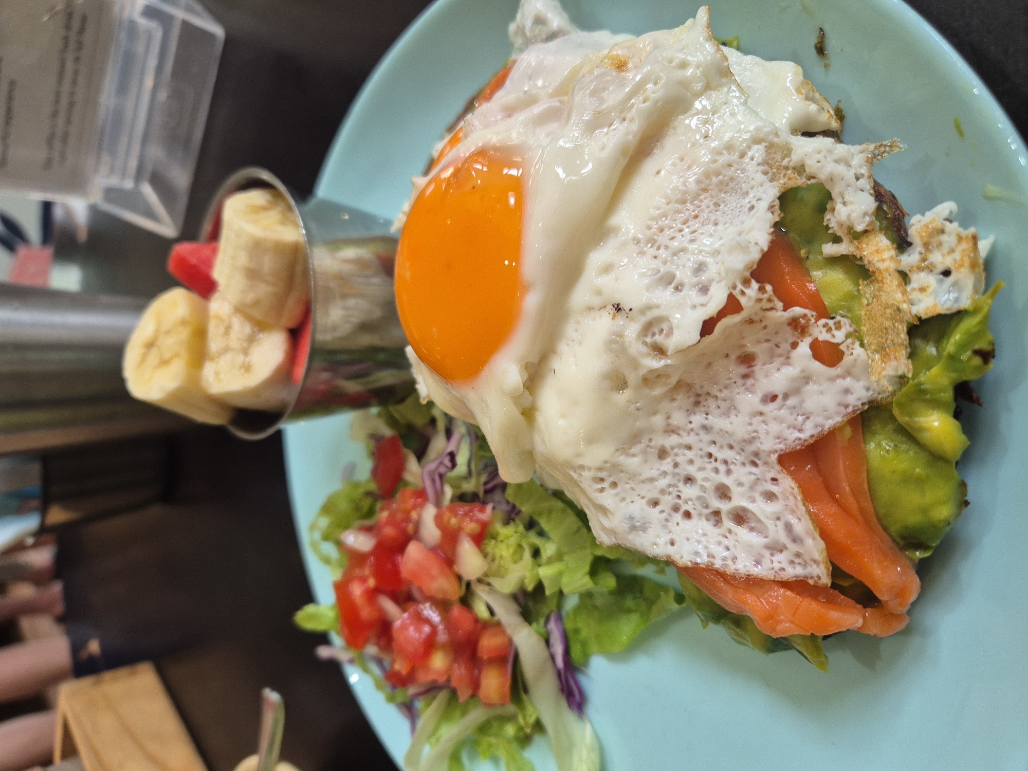 A plate of food rests on a dark surface. The plate is a light blue and contains a breakfast dish. The dish consists of a piece of toast topped with mashed avocado, slices of smoked salmon, and a fried egg with a bright orange yolk. To the left of the main dish is a small salad of lettuce, chopped tomatoes, and red onion. In the background, a metal cup holds slices of banana and watermelon. The scene appears to be in an indoor setting, possibly a cafe, with out-of-focus figures and objects in the background, suggesting a public space.