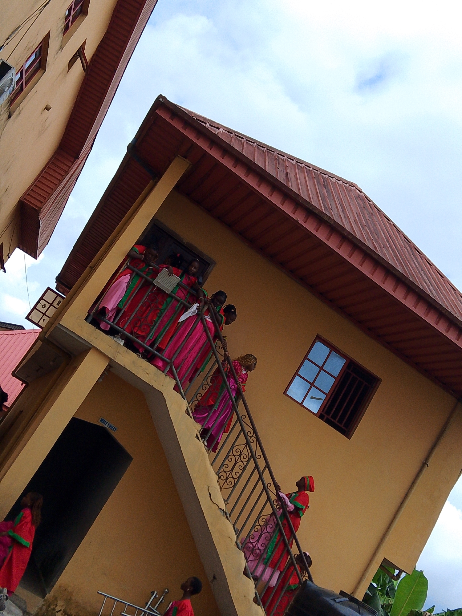 This media file depicts an exterior shot of a two-story building with yellow walls and red corrugated roofing. The building has an open staircase with ornate metal railings leading up to the second floor. Several people, predominantly women and girls dressed in vibrant pink and red traditional attire with green accents, are visible on the staircase and on a balcony on the second floor. Some are looking down, while others are engaged in conversation or looking towards the camera. A sign above a dark doorway on the first floor reads "MALE TOILET."
The scene appears to be set during the daytime, under a cloudy sky, suggesting it might be an overcast day. The presence of traditional clothing and the architectural style, combined with the "Male Toilet" sign, hint at a location in Nigeria, specifically Yenegoa as per the context provided. The people's expressions are neutral or slightly smiling, suggesting a calm and possibly festive atmosphere. The building's design, with its external staircase and prominent roofing, is characteristic of some residential or community buildings in the region. The image is taken from a low angle, looking upwards towards the building and the sky.