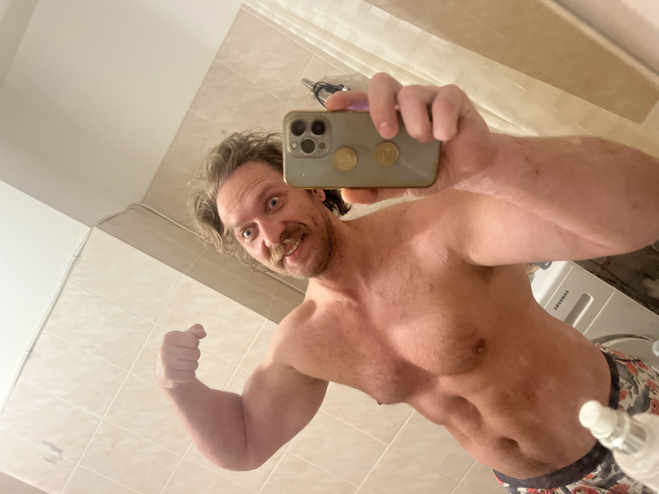A shirtless man poses for a bathroom selfie. He flexes his left arm, showcasing his bicep. His face is close to the camera, displaying a wide-eyed expression and a mustache. The man is holding a smartphone with two coins glued to the back. In the background, light beige tiles cover the walls, a white washing machine is visible, and a small white bottle sits on the counter.