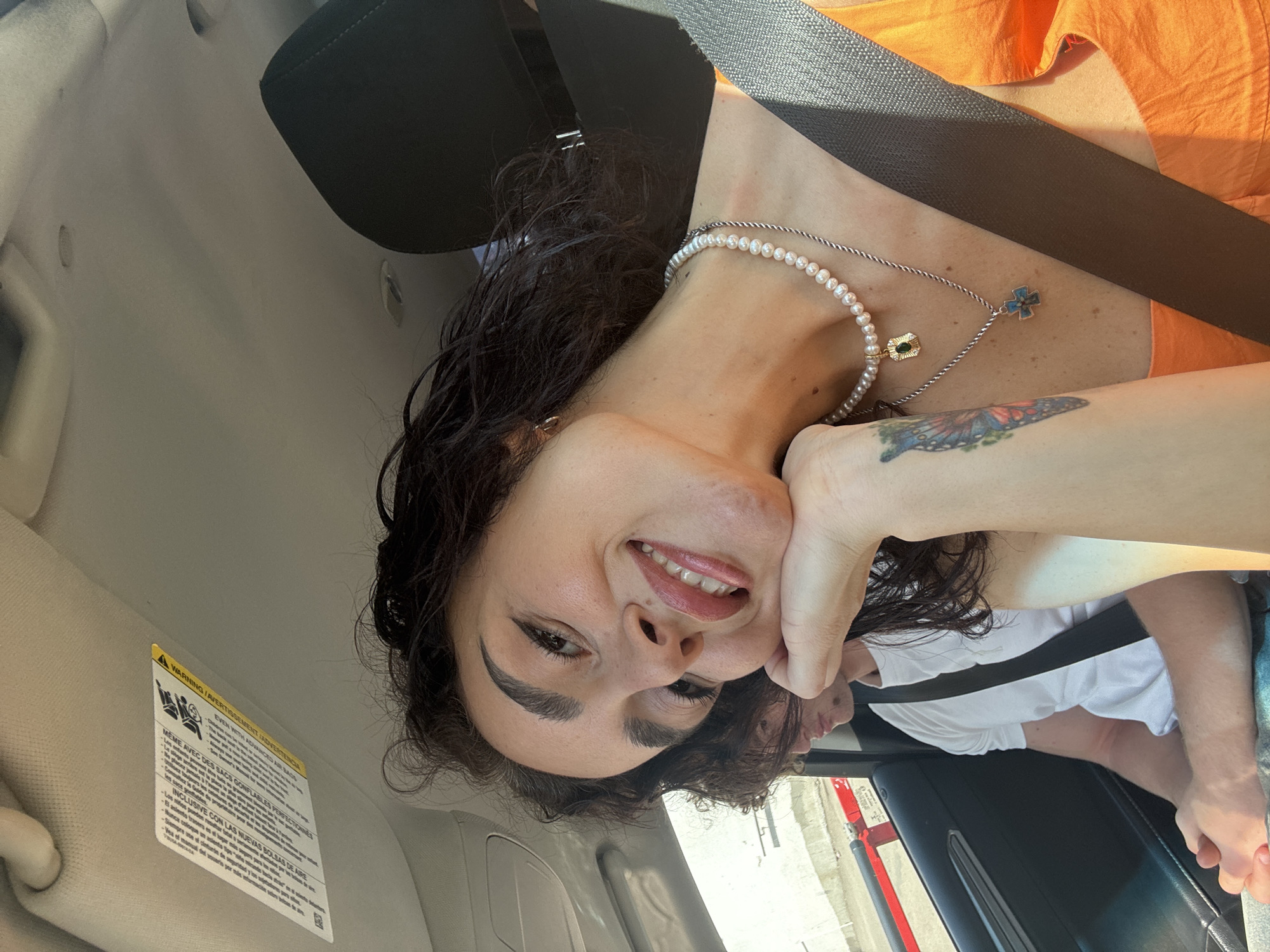 A woman with dark, curly hair is in the passenger seat of a car, smiling as she rests her chin on her hand. She is wearing an orange top, a pearl necklace, and a silver necklace with a cross pendant. A butterfly tattoo is visible on her forearm. In the background, a man with light brown hair wearing a white t-shirt is visible in the driver's seat, with a seatbelt across his chest. The car interior, including the dashboard and sun visor with a warning label, is visible. Outside the car window, a blurry view of a city street with a red barrier and buildings can be seen. The lighting suggests it is daytime.