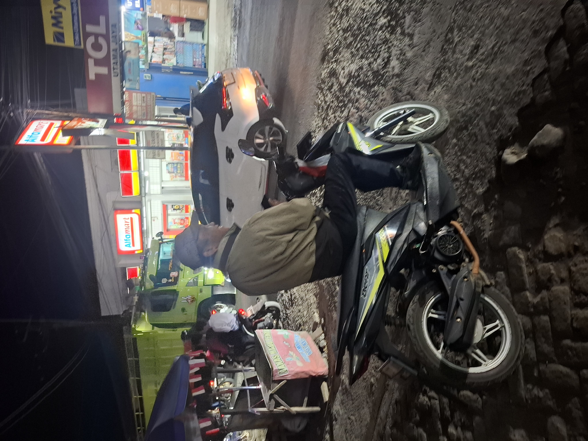 The image depicts a nighttime street scene in Ciheulang Tonggoh, Indonesia. A man wearing a brown jacket and a cap is seated on a black and yellow scooter, facing away from the camera. He is wearing dark pants and black shoes. The scooter is parked on a rough, uneven surface that appears to be a dirt road or ground. In the background, a bright green truck is visible, as well as a white car with its brake lights illuminated. Adjacent to the truck, a building with red and white signage reads "Alfamart". Another sign above it displays the "Alfamart" logo. Further to the right, a sign with "TCL" branding is prominent. Several other smaller signs and store entrances are visible. A blue awning or shelter is present to the left of the scooter. The overall environment suggests a commercial area with shops and busy traffic, even at night.