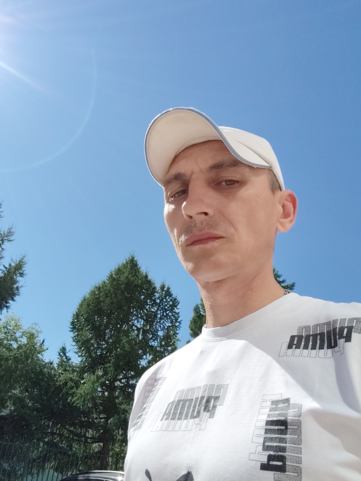This is a close-up, low-angle selfie of a man wearing a white baseball cap and a white t-shirt with a repeating black and grey graphic print. He has short brown hair, brown eyes, and a thin mustache. The man is looking directly at the camera with a neutral expression.
The background is a clear, bright blue sky with some lens flare from the sun in the upper left corner. Below the sky, there are dark green evergreen trees, suggesting an outdoor setting, likely in a natural or park-like environment. The presence of trees and the bright daylight indicate it is daytime, likely sunny.
The visible text on the t-shirt appears to be a stylized graphic that repeats multiple times. While the exact wording is difficult to decipher completely, it seems to be an abstract or branded word.
The image was taken in Bor, Russia, based on the provided location context. The bright sun and blue sky suggest pleasant weather conditions. The overall mood of the image is casual and straightforward.