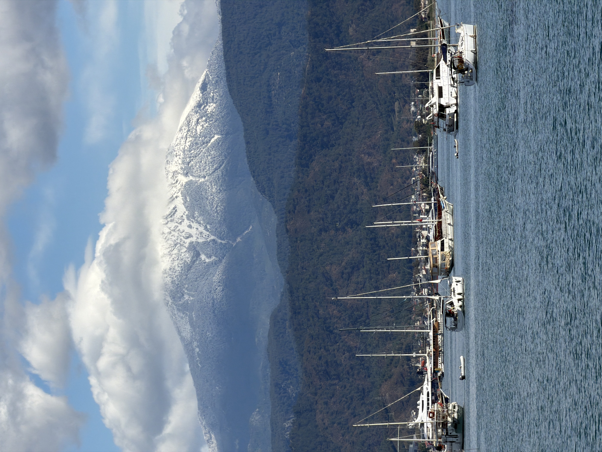 The image depicts a view in Fethiye, Turkey, featuring a line of sailboats docked in a harbor. The water occupies a significant portion of the frame on the right side. The boats are varied in size and design. Their masts point upwards, creating a linear pattern.
Behind the boats is a lush, green mountain range which occupies the middle-ground. To the left of the mountains, a snow-capped peak is partially obscured by clouds. The sky above the mountains is a mix of blue patches and white clouds.