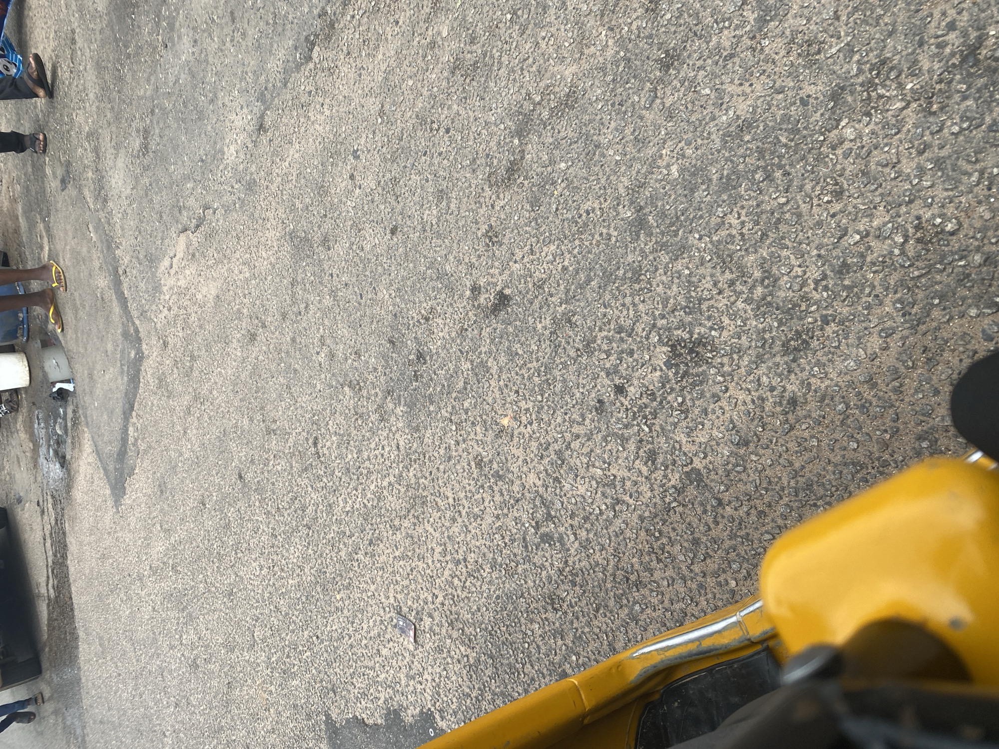 The image shows a ground-level view of a paved area, possibly a street or walkway, with scattered debris. In the upper left portion of the frame, several people are partially visible, wearing casual clothing and footwear such as flip-flops. Their legs and feet are the primary focus in this area. To the right, a bright yellow object, likely a vehicle or part of a vehicle, is partially in view, dominating the lower right side of the image. The ground itself is a rough, grey asphalt surface with small stones and some darker patches, suggesting it might be wet or dirty in places. There are no clear indicators of the time of day or specific weather conditions, though the lighting suggests it is daytime. The scene appears to be an outdoor urban or semi-urban environment, possibly in Jalingo, Nigeria, as indicated by the location context, though no specific landmarks are visible. The overall impression is one of everyday street activity.