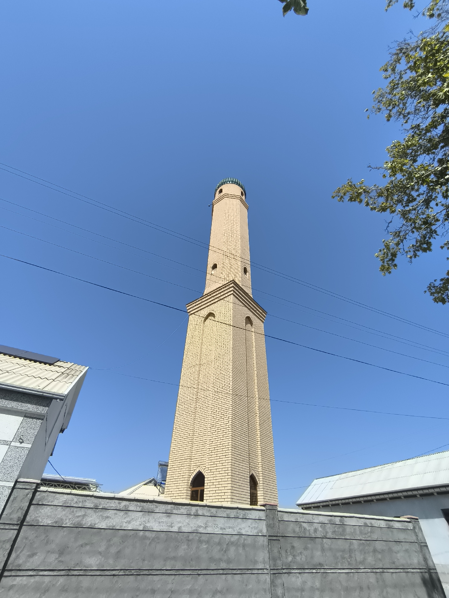 A tall, slender minaret, constructed from light yellow bricks, dominates the center of the image, reaching towards a clear blue sky. The minaret has a ribbed, green dome at its apex and features several arched openings and decorative brickwork along its height. In the foreground, a grey concrete wall runs horizontally across the bottom of the frame, partially obscuring the base of the minaret. To the left of the minaret, the corner of a building with a white, corrugated roof is visible, juxtaposed with a section of a grey and white patterned wall. To the right of the minaret, another building with a white, sloped roof can be seen. Several dark electrical wires stretch across the blue sky, traversing the scene from left to right. A cluster of green leaves from a tree branch hangs from the upper right corner of the image. The overall scene depicts an urban environment, likely within the city of Babakhodja, Uzbekistan, with a prominent religious structure.