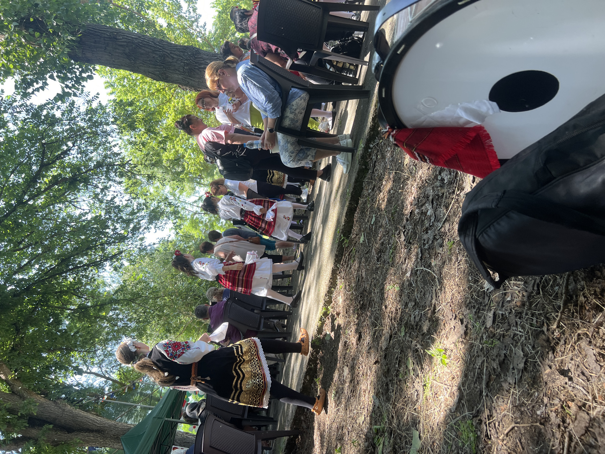 This image captures an outdoor cultural gathering on a clear, sunny day in Bolyarovo, Bulgaria. The upper portion of the frame is dominated by the dense, green foliage of large deciduous trees, with bright sunlight filtering through, casting dappled shadows across the scene.
In the mid-ground, a paved pathway separates a dirt-covered foreground from an assembly of people. Several individuals are dressed in traditional Bulgarian folk costumes. A woman in the left-center, viewed from behind, wears a black vest over a white blouse featuring red, white, and green embroidery on the sleeves, secured by a brown woven belt. Her black skirt displays gold-colored wavy patterns along the hem, worn over a white lace-trimmed petticoat. She wears black tights and light brown, traditional-style footwear, and her hair is in a ponytail adorned with white flowers. She holds a piece of white fabric. To her right, another individual in a similar costume, likely a younger female, is partially visible from behind, wearing a white embroidered blouse, a red and white striped skirt, and black shoes.
Numerous attendees are present, some seated on dark plastic chairs on the paved area, and others standing. A woman in the right-center is seated, wearing a light blue striped shirt, patterned capri pants, and white athletic shoes, holding a plastic water bottle. Other individuals are dressed in casual clothing. On the far left, a green tent or canopy structure is partially visible.
The foreground consists of dark, uneven soil covered with dry leaves, small twigs, and sparse green plant growth. In the lower right corner, a black soft-sided bag rests next to a large, white drum (likely a tapan) with a prominent black circular accent. A red and white striped woven fabric is draped over the rim of the drum.