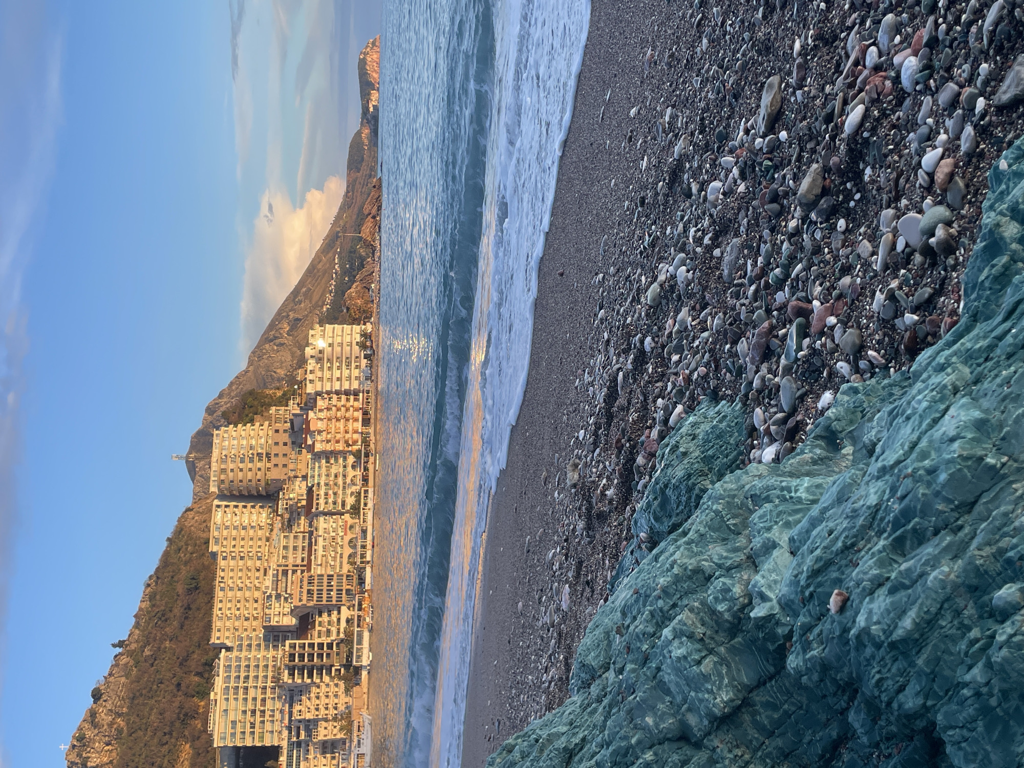 The image displays a coastal landscape in Rafailovići, Montenegro, captured during golden hour. In the foreground, a large, angular rock formation exhibiting a greenish-blue coloration occupies the lower left. To its right, a pebble beach extends, composed of numerous small, smooth stones in shades of grey, white, brown, and some reddish or teal. Waves are actively breaking on the shore, creating white foam at the water's edge.
The mid-ground features the sea, which has a prominent turquoise hue and moderate wave action. Reflections of warm, golden-orange light are visible on the water's surface, particularly near the shore.
The background presents the town of Rafailovići, densely packed with multi-story buildings, primarily white or light-colored, constructed into a steep, rocky, and partially vegetated mountainside. These structures, likely hotels or apartments, feature numerous windows and balconies, all illuminated by the warm, low-angle sunlight. A white cross is visible atop the highest peak of the mountain range to the far left, above the town. Further to the right along the coastline, additional rugged mountains and a distinct reddish-brown rocky headland project into the sea. The sky above is a clear blue, with scattered white and light pink-tinted clouds, particularly towards the right horizon. No individuals or specific human activities are discernable within the scene. Several small vessels are moored near the waterfront buildings.