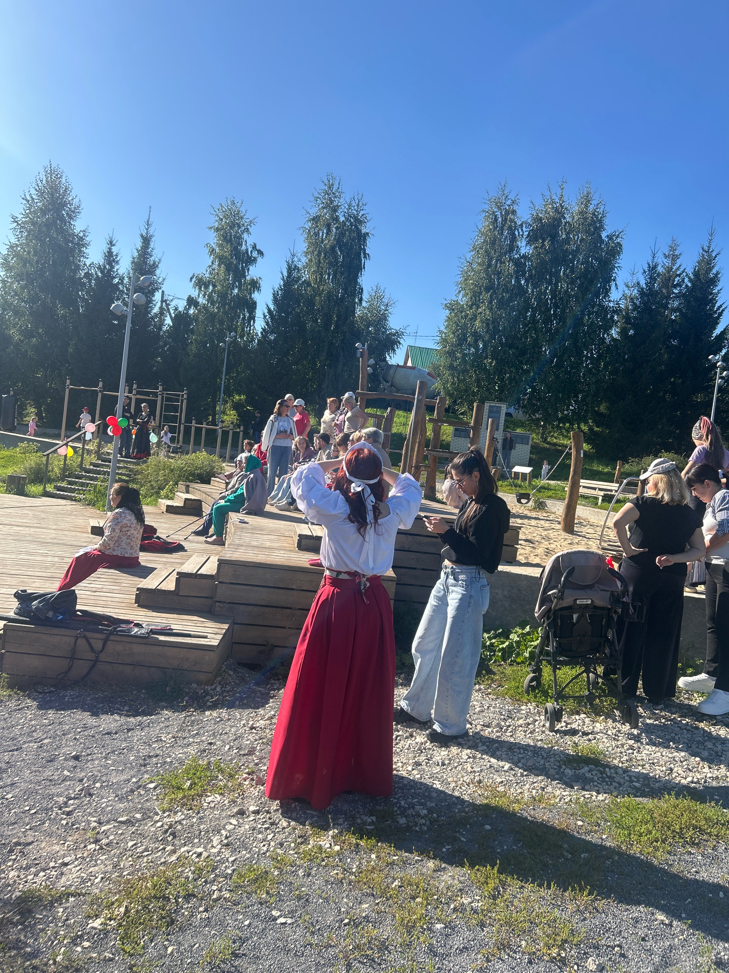 In an outdoor public space, possibly a park or community event area, a group of people are gathered under a clear blue sky. The terrain is mixed with gravel and patches of grass. In the foreground, a person in a white blouse and a long, flowing red skirt stands with their hands on their head, wearing a white blindfold or headband. To their left, another person in a white and patterned top and red pants is seated on a wooden structure. Behind them, several other individuals are visible, some sitting on wooden risers, and others standing. A playground with wooden structures and a sandpit is present in the background. Trees line the perimeter of the area, with streetlights interspersed among them. A stroller is parked on the gravel. The scene appears to be a daytime event, possibly a performance or cultural gathering.